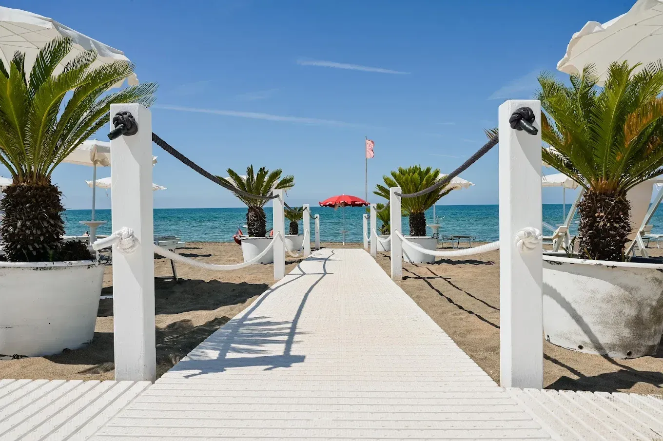 Una passerella bianca conduce a una spiaggia con palme, ombrelloni e l'oceano blu sotto un cielo limpido.