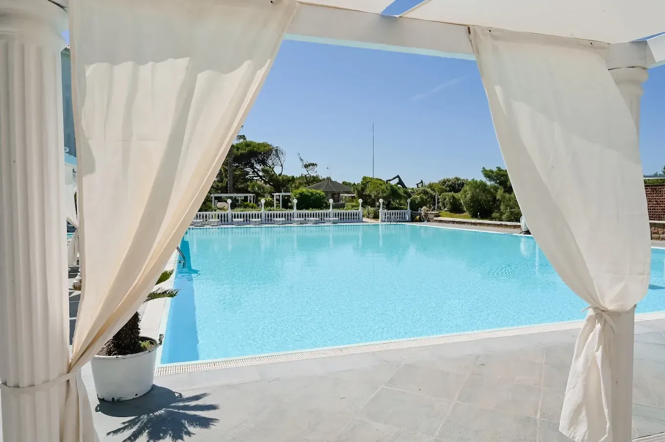 Delle tende bianche incorniciano una piscina blu brillante del resort, con lettini e alberi sullo sfondo.