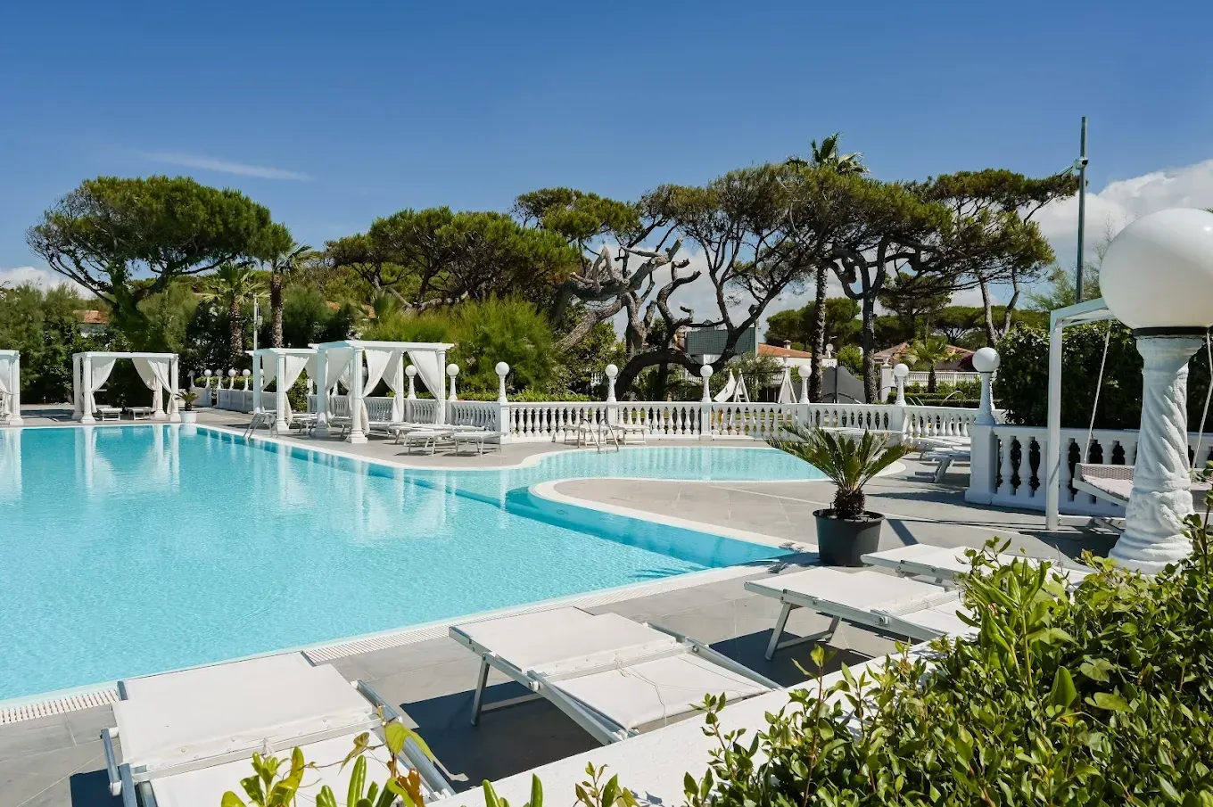 Piscina all'aperto con acqua turchese, lettini bianchi e pergolati bianchi sotto i pini in una giornata di sole.