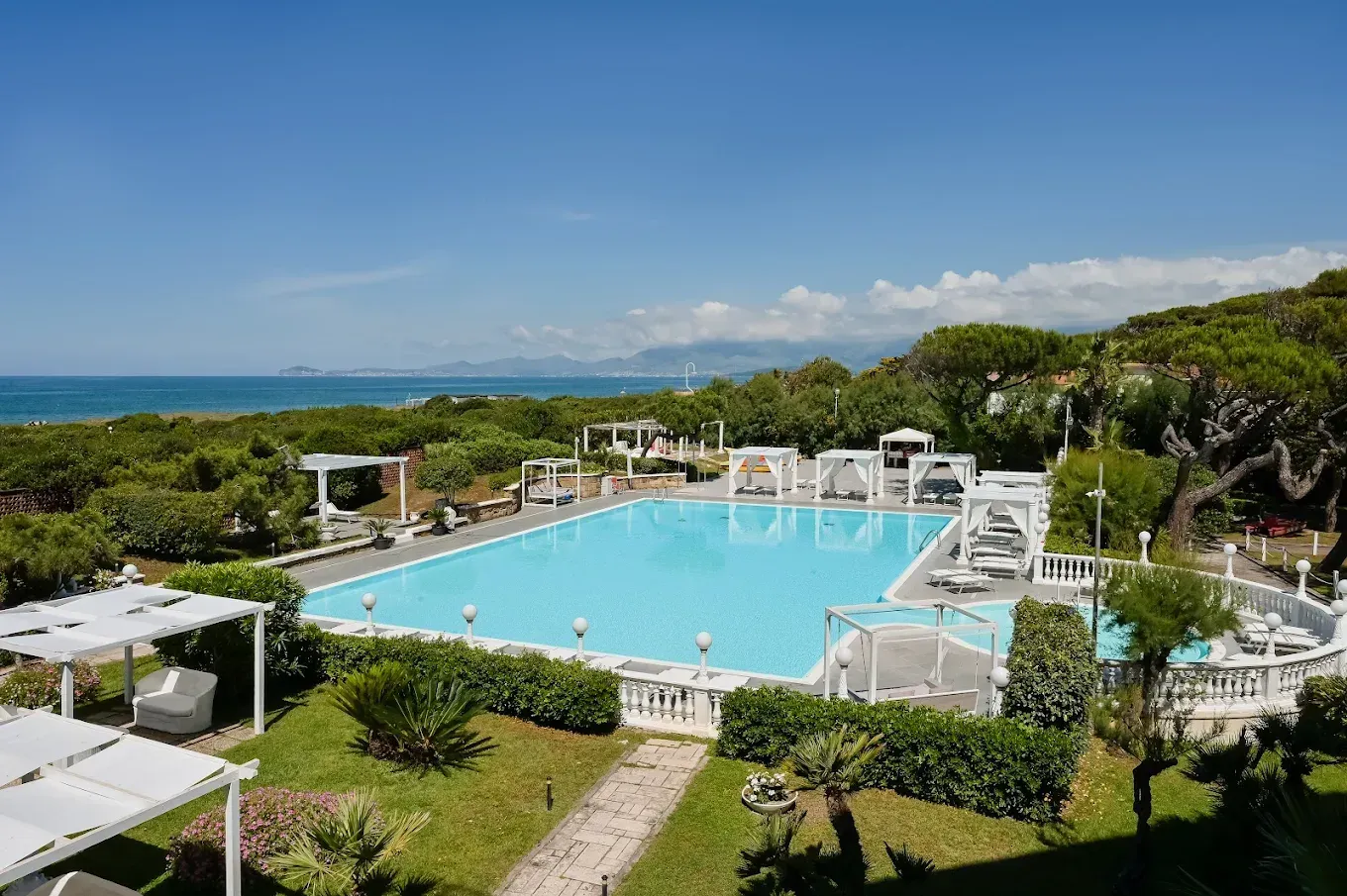 Piscina del resort con vista sull'oceano, cabine bianche e vegetazione lussureggiante sotto un cielo azzurro.