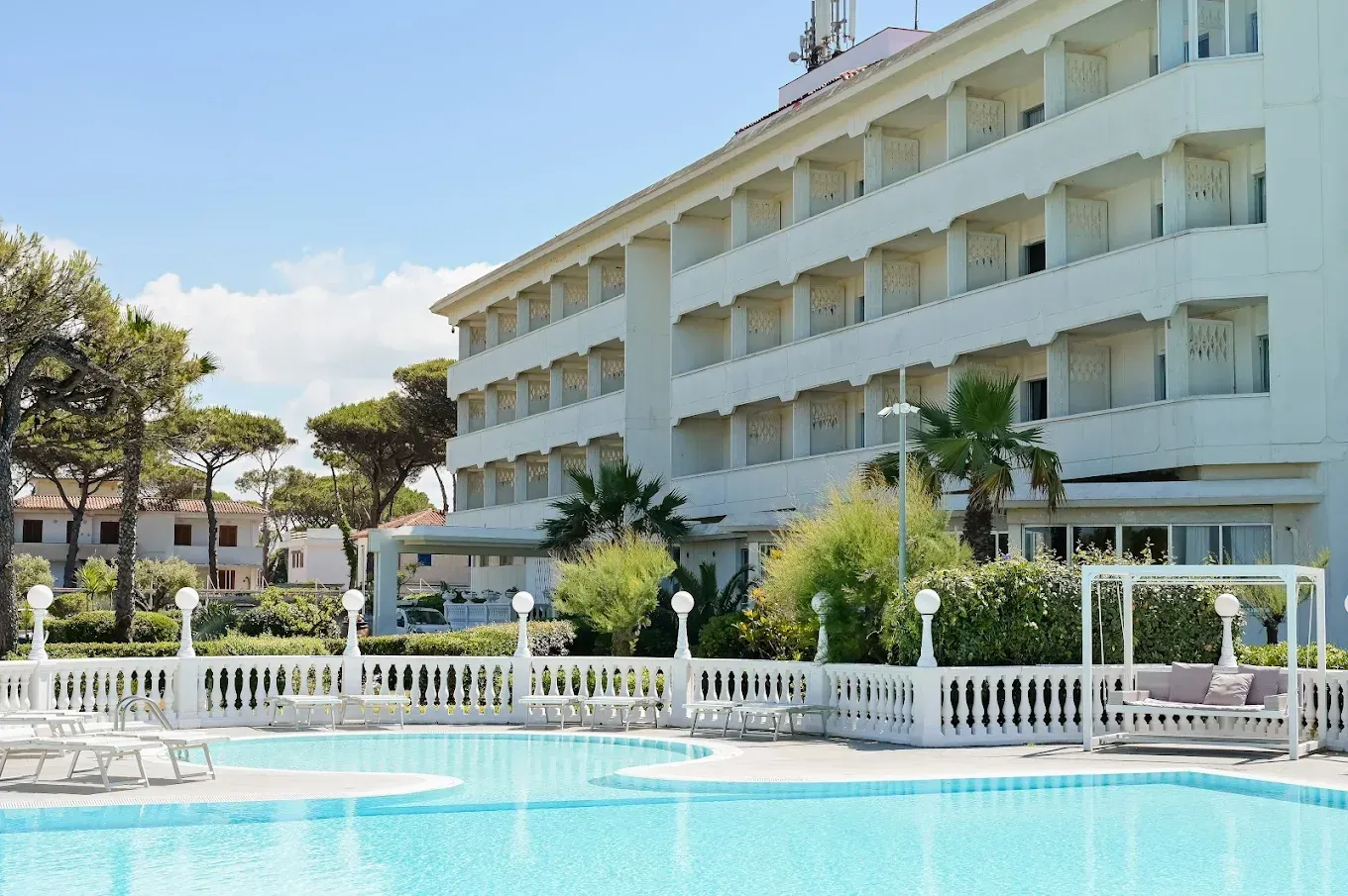 Un edificio alberghiero bianco accanto a una piscina turchese con una recinzione bianca e palme sotto un cielo azzurro limpido.