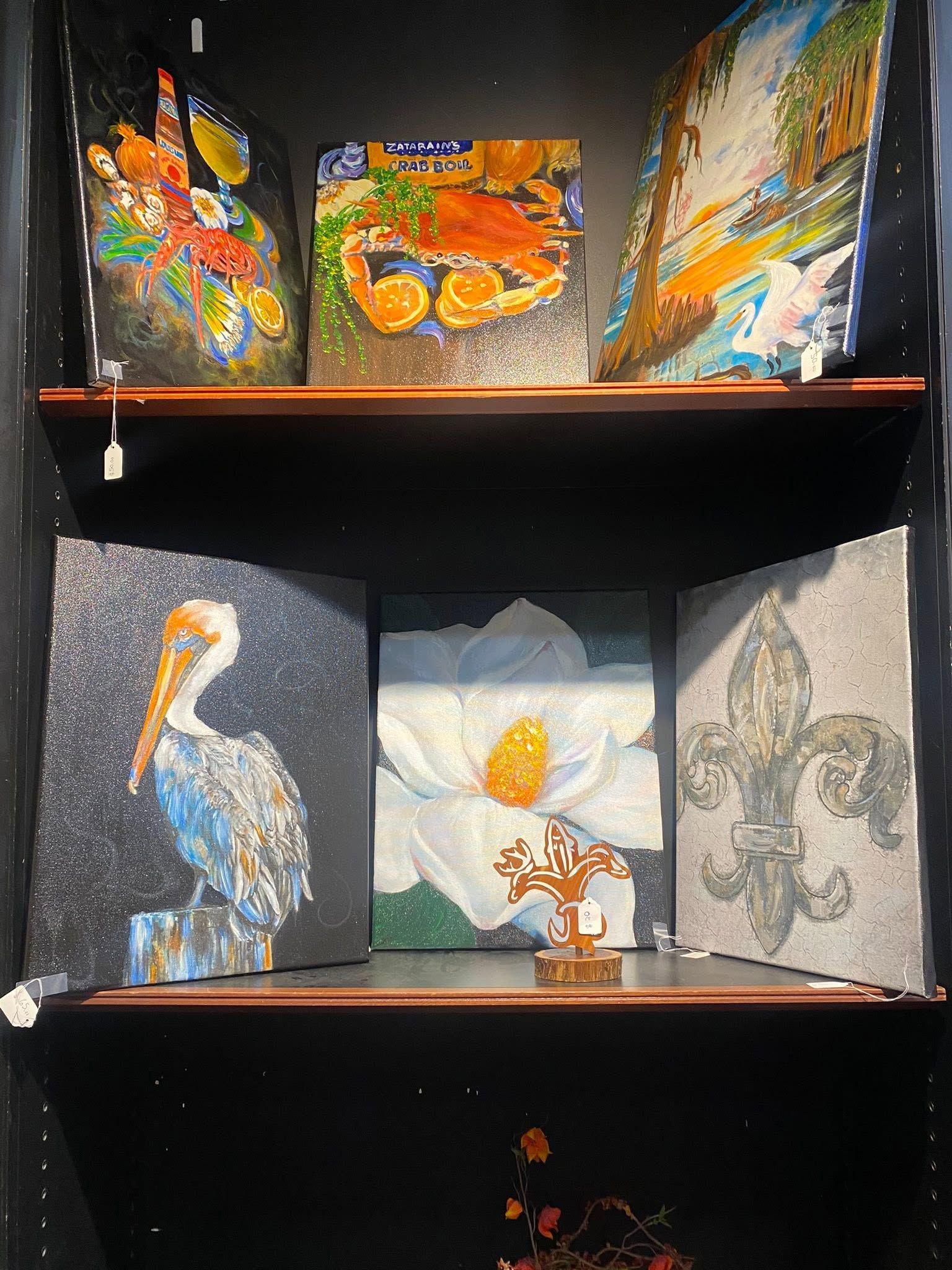 Artwork display on a black shelf, with colorful bird, flower, and abstract paintings.