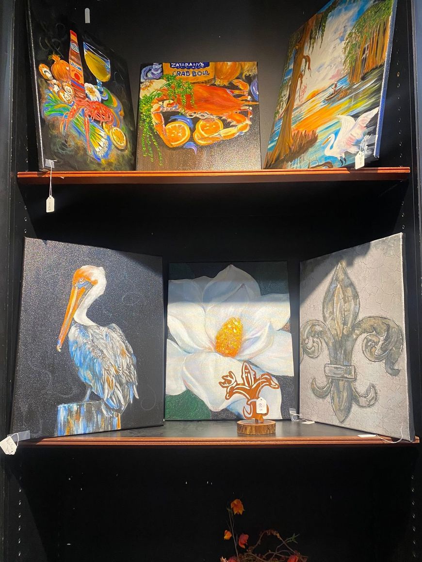 Artwork displayed on a black shelving unit: colorful panels above and bird, flower, and fleur-de-lis paintings below