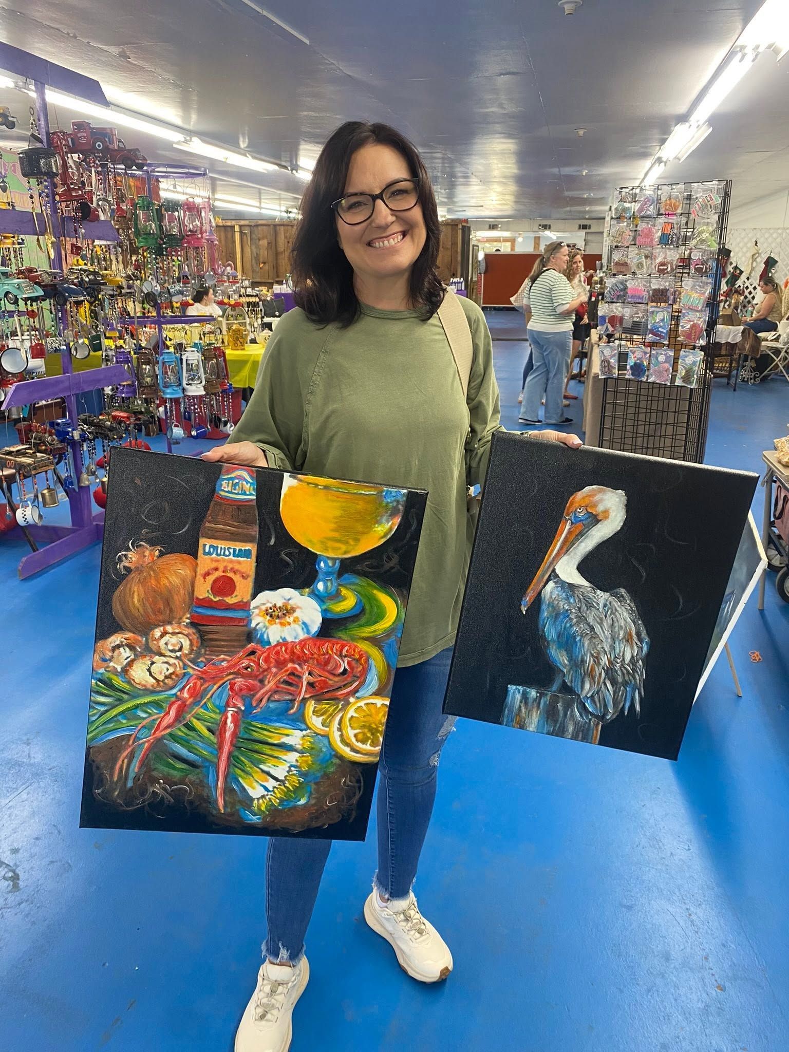 Woman in a market aisle holding two colorful paintings, including a bird and a cocktail.