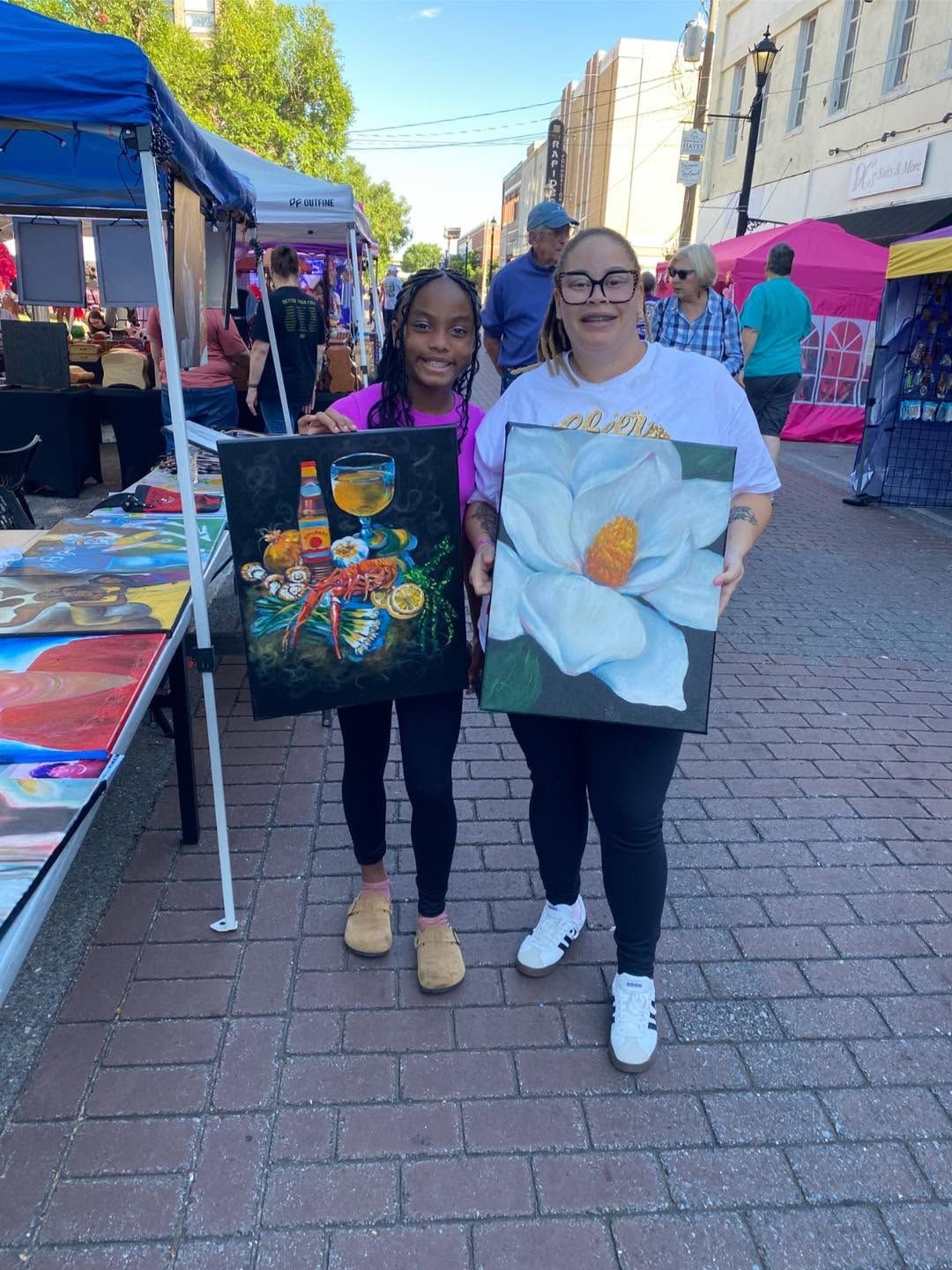 Two people holding flower paintings at an outdoor market stall on a sunny street