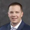 Studio headshot of a person in a navy suit and patterned tie against a gray backdrop