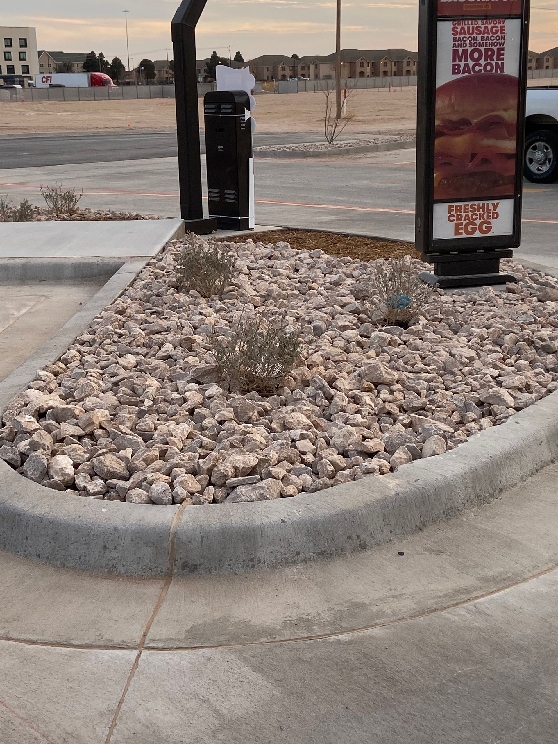 Rock mulch landscaping bed beside a drive-thru menu kiosk in a parking lot.