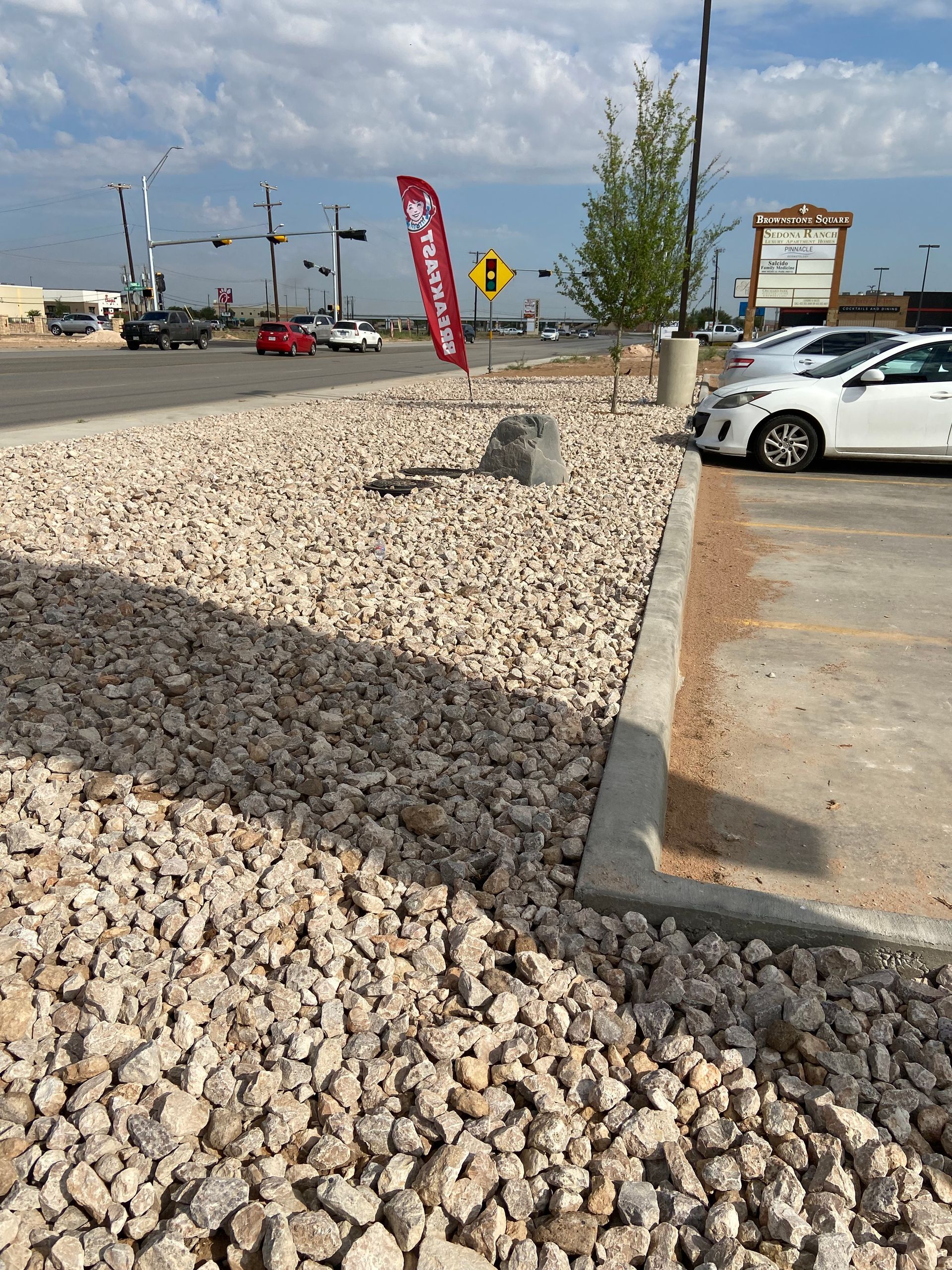 Rock-covered parking lot edge beside white cars, red flag sign, and cloudy sky near an intersection.