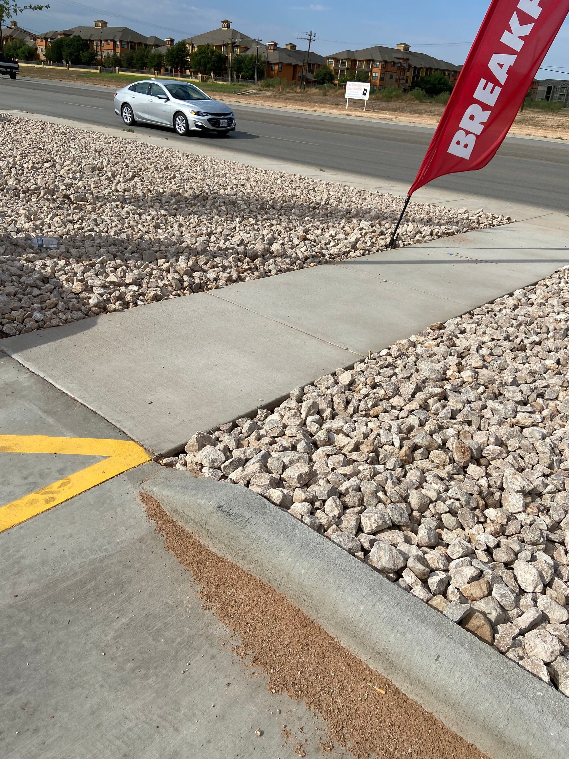 Car driving on a gravel lot beside a concrete curb and red flag banner under a clear sky