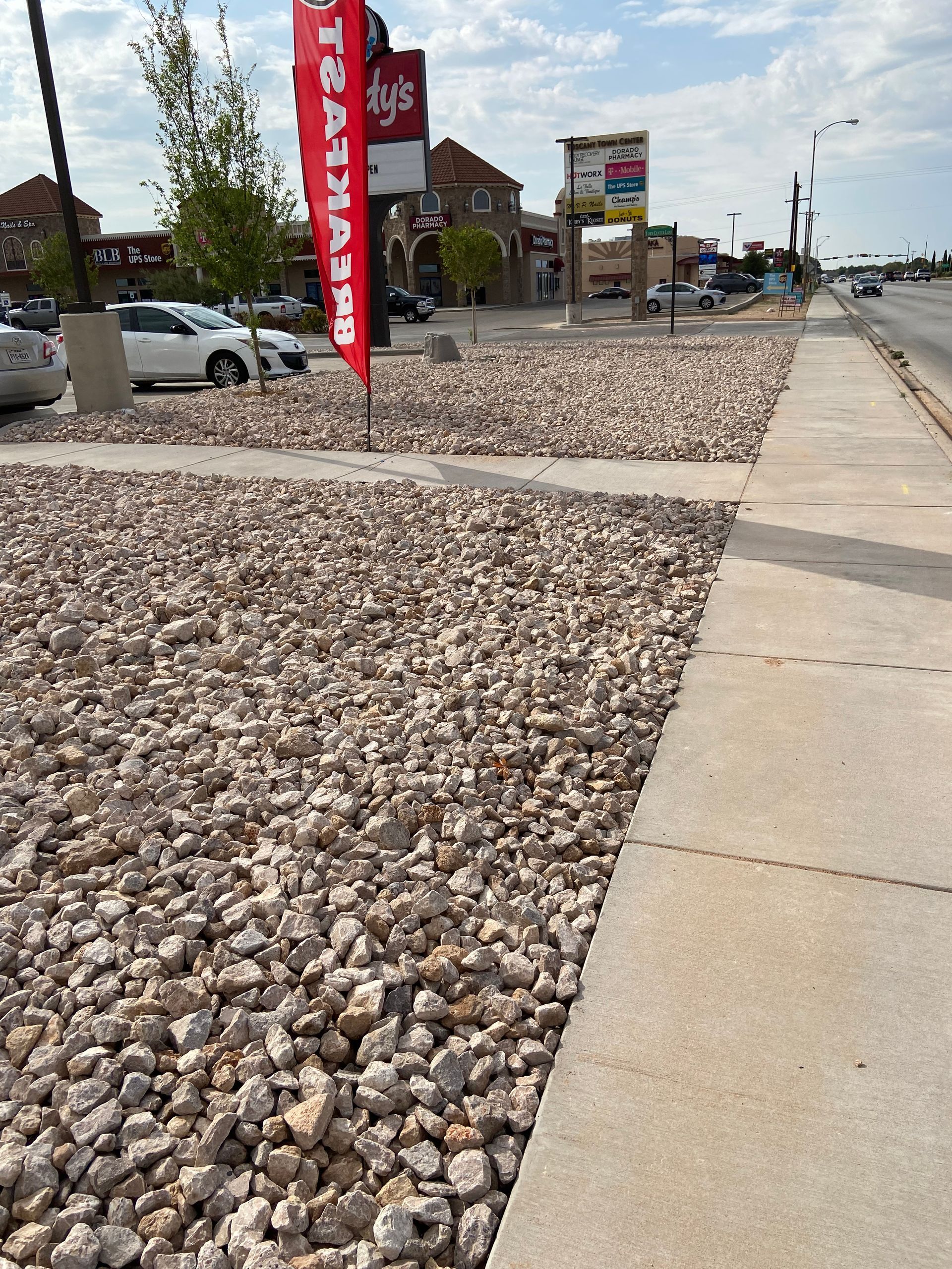 Rock-covered roadside beside a sidewalk, with storefront signs and parked cars under a clear sky.
