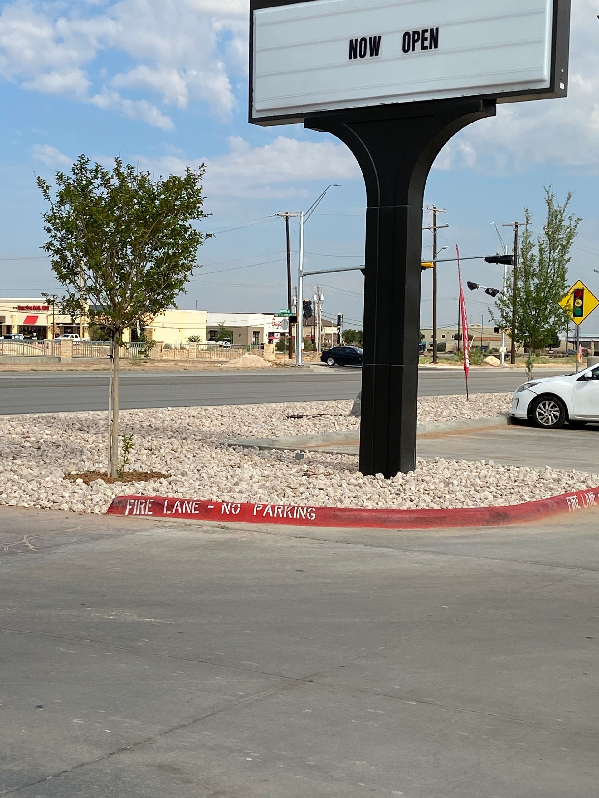 Empty roadside sign on a gravel median with a small tree and a white car in the background.
