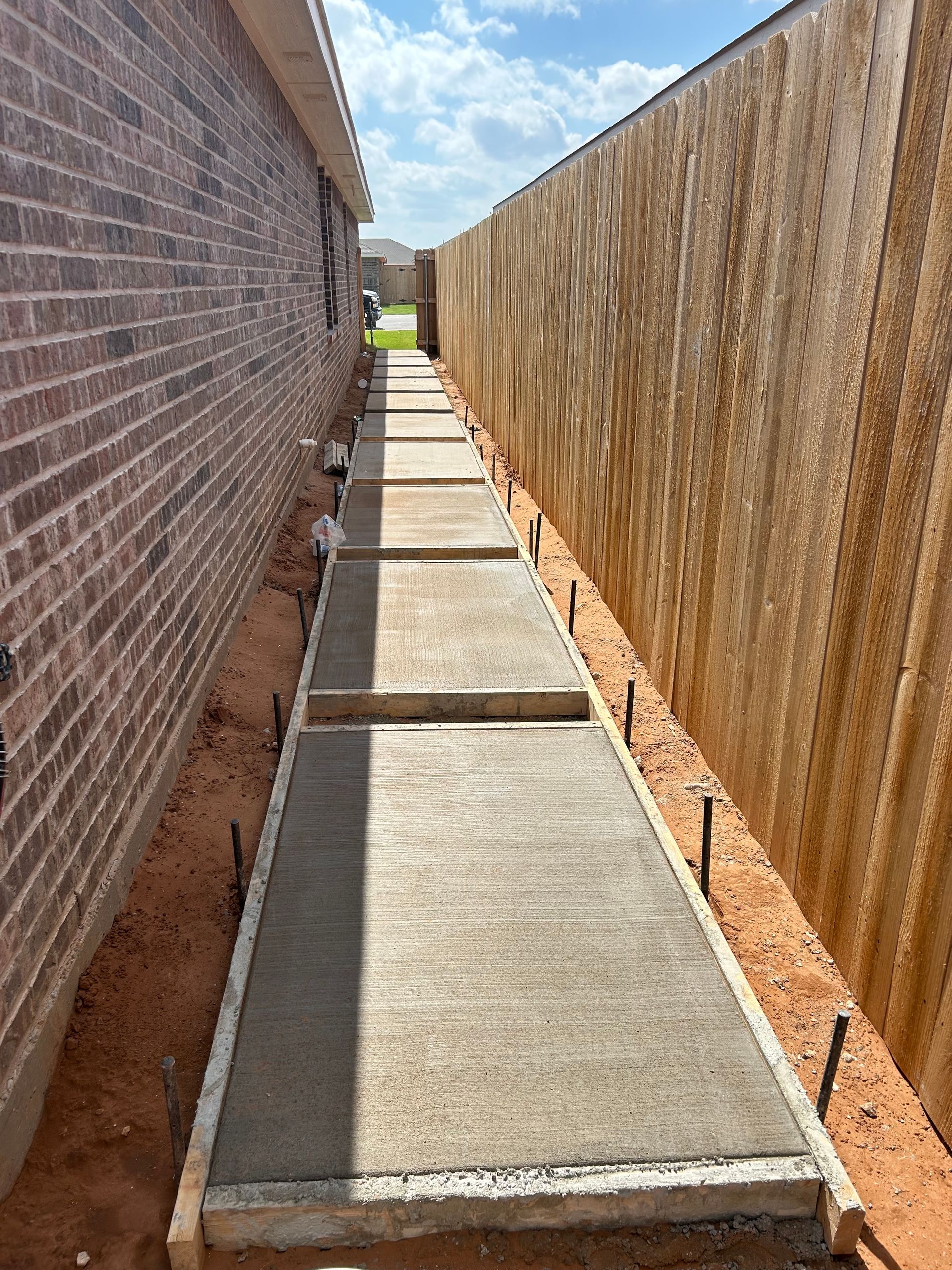 Narrow concrete walkway between brick wall and wooden fence, leading to a bright opening ahead
