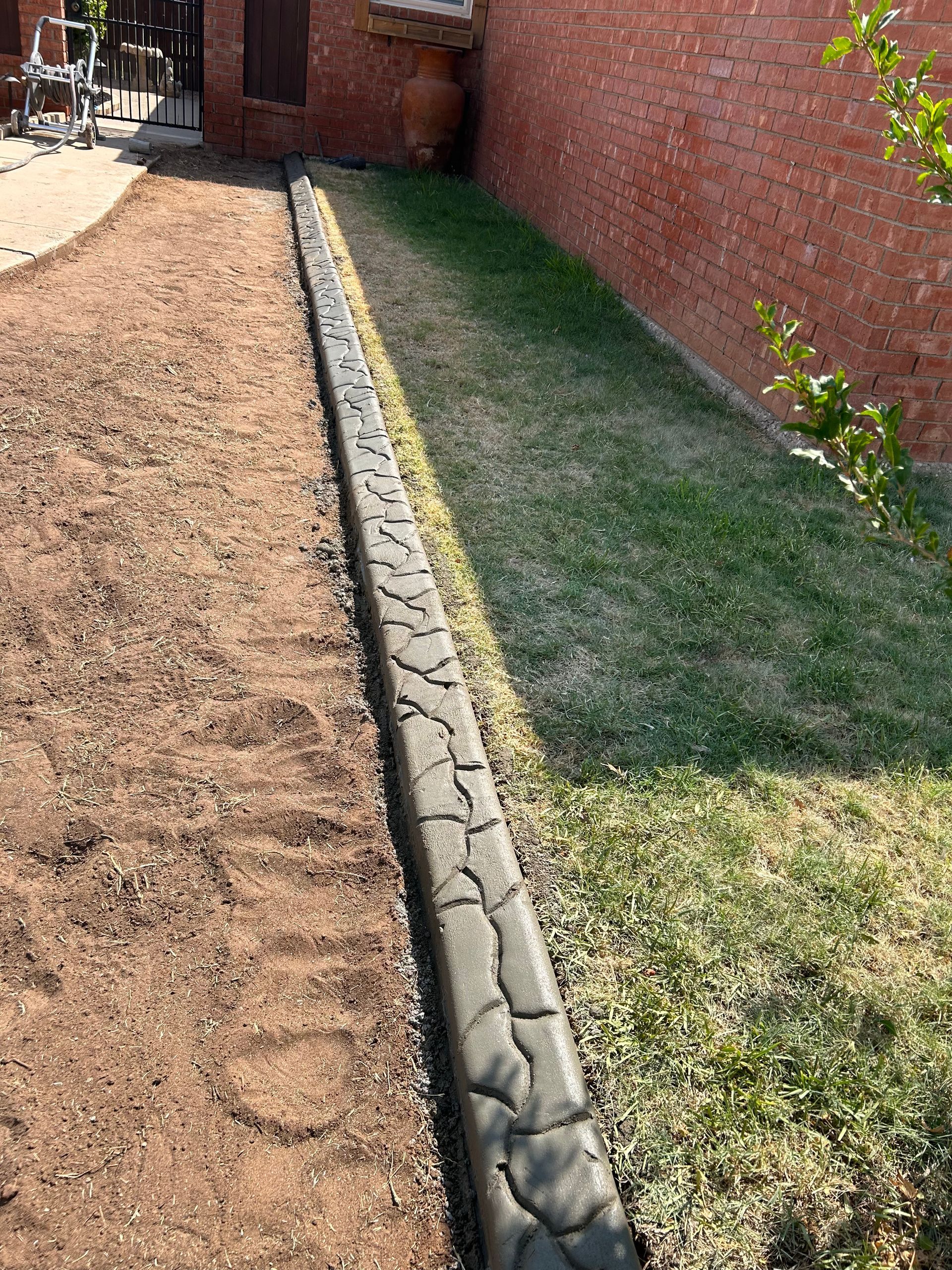 Narrow concrete border divides brown mulch and green grass beside a brick wall and walkway.