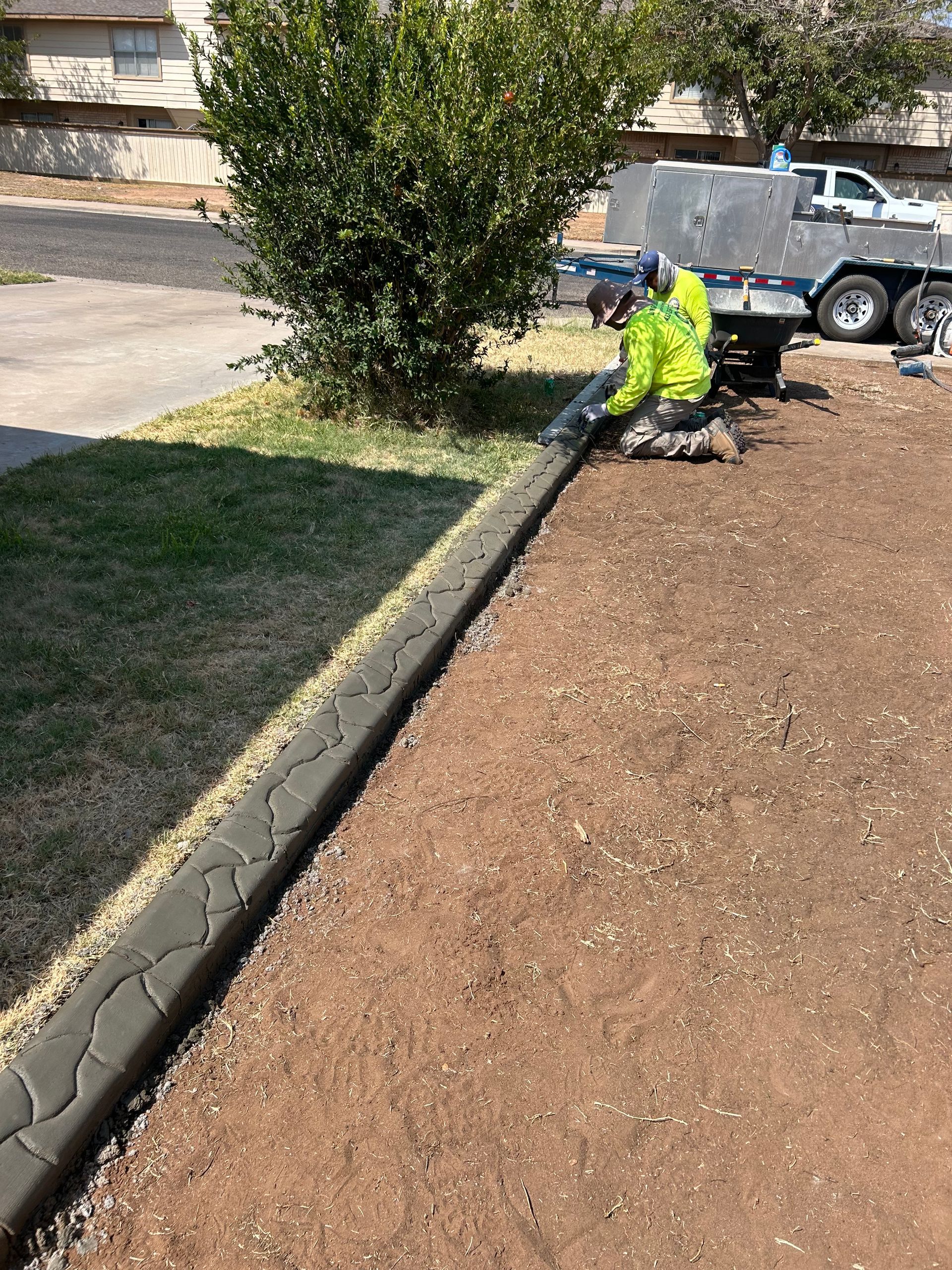 Worker in neon safety vest edging a curb beside a dirt lot and driveway.
