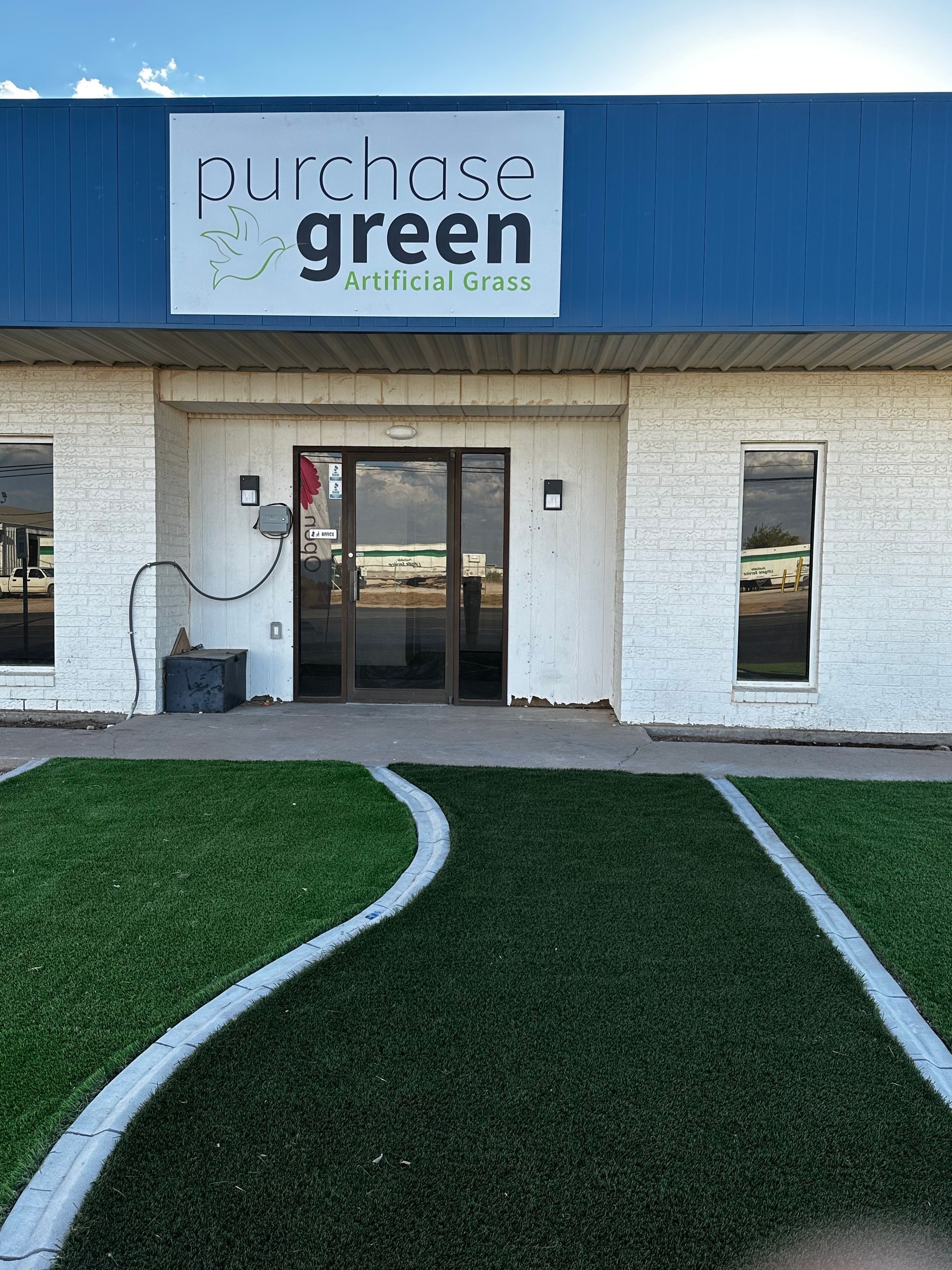 Storefront with a “purchases green” sign above a glass door and artificial grass walkway leading to the entrance.