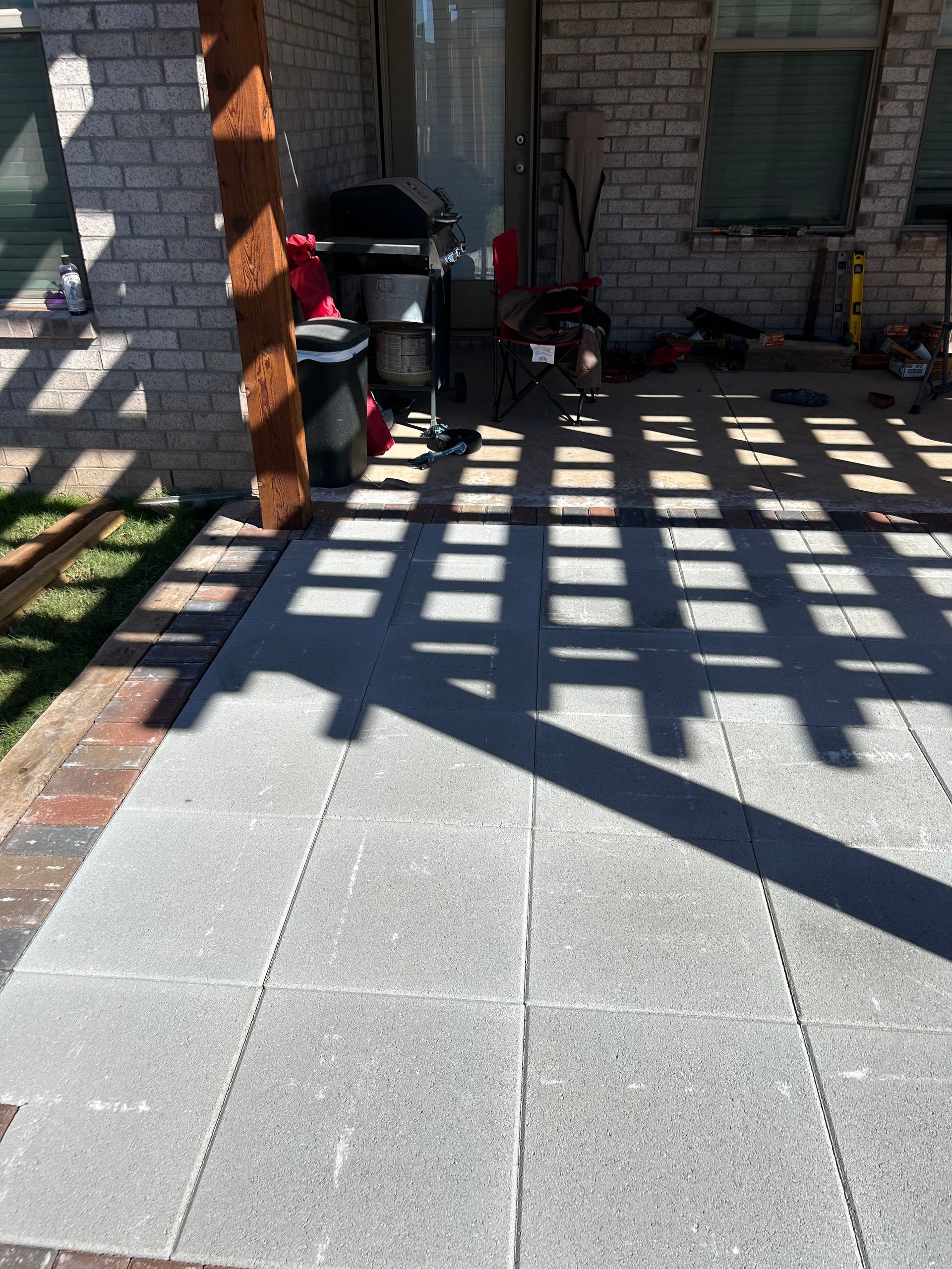 Sunlit patio with shadowed railing pattern, brick wall, grill, and potted plants by a doorway