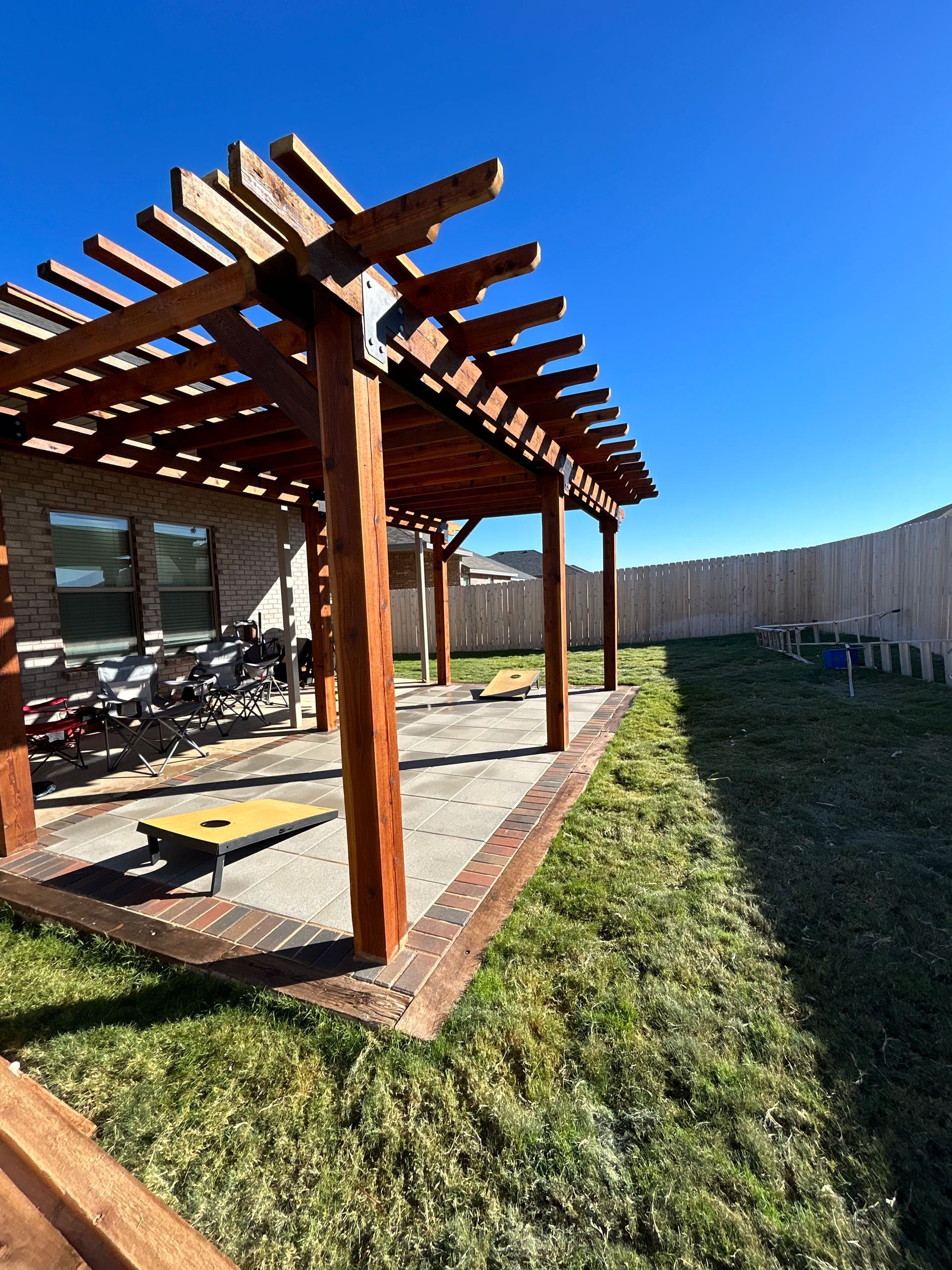 Wooden pergola beside a patio on a sunny backyard lawn, with a fence and blue sky.