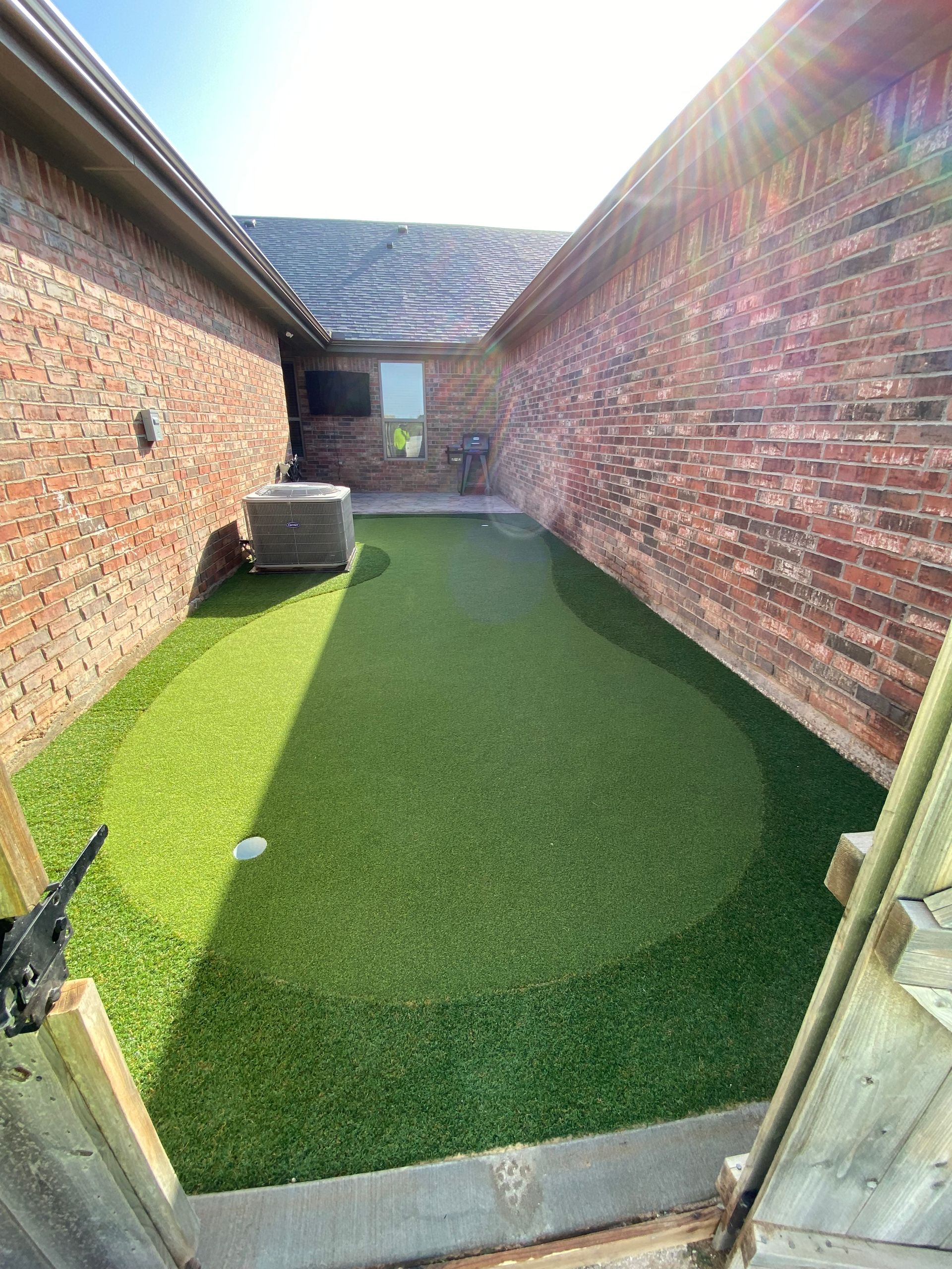 Narrow brick courtyard with artificial turf and a small round planter under an open sky.