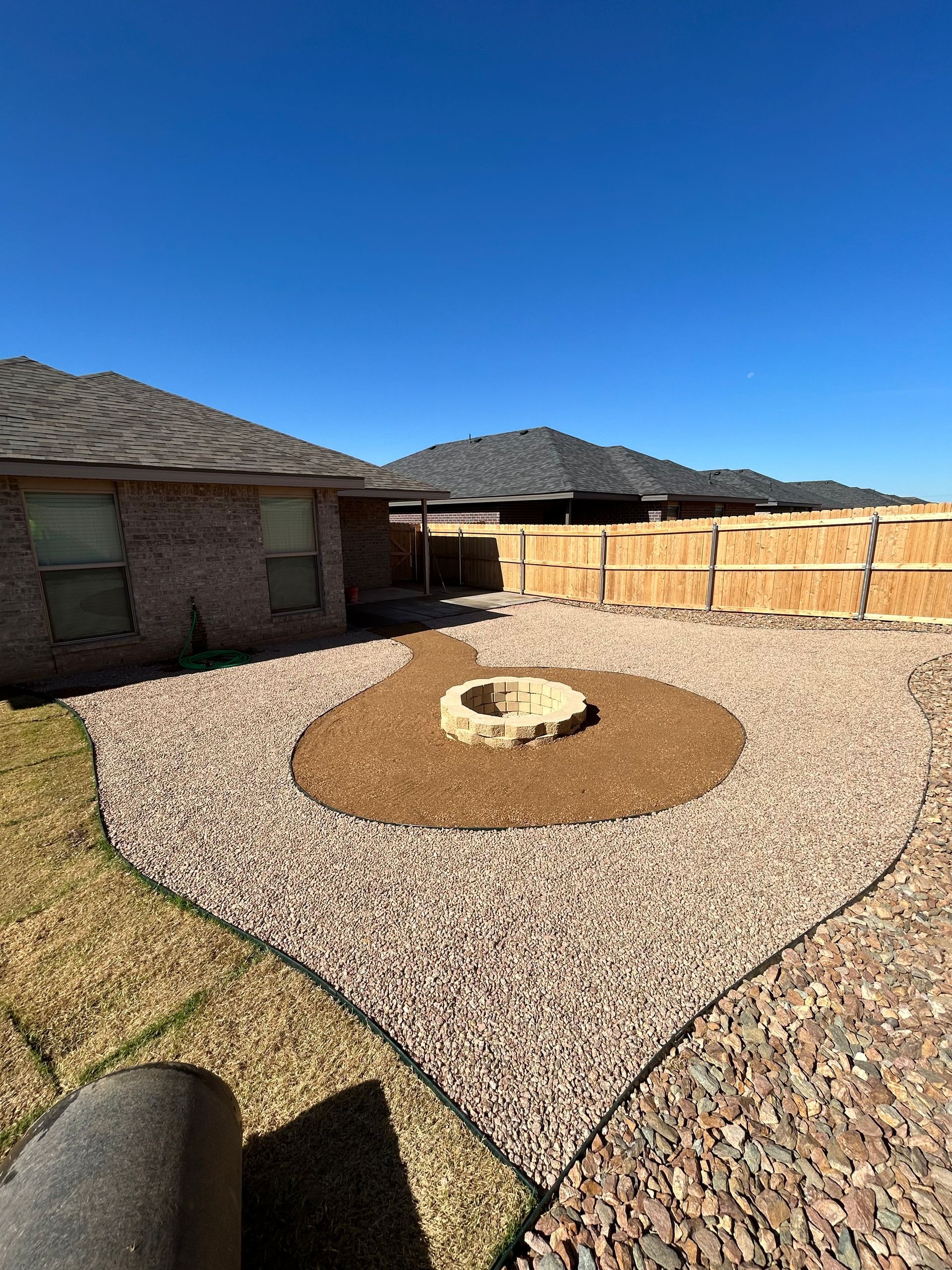 Backyard patio with gravel, circular fire pit, wooden fence, and blue sky