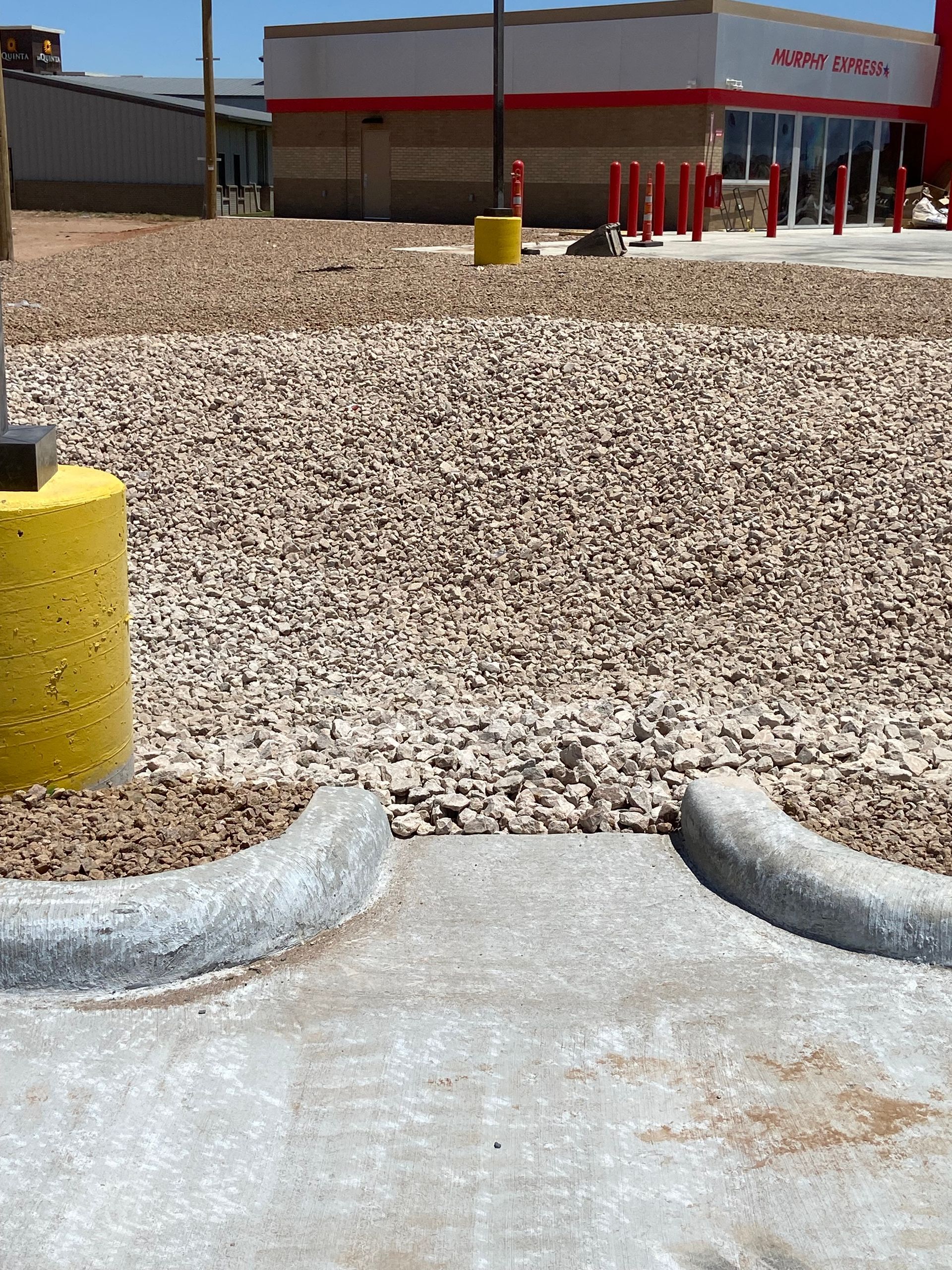 Gravel lot outside a storefront with red trim, yellow bollard, and concrete curb in the foreground