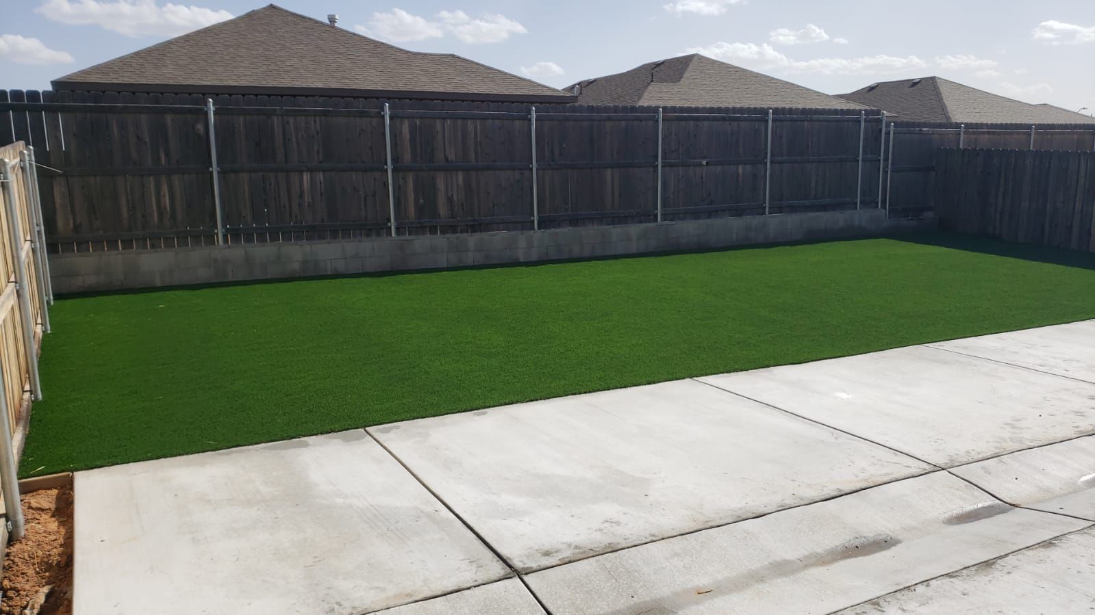 Backyard with green artificial turf, concrete patio, and wooden privacy fence under sunny skies