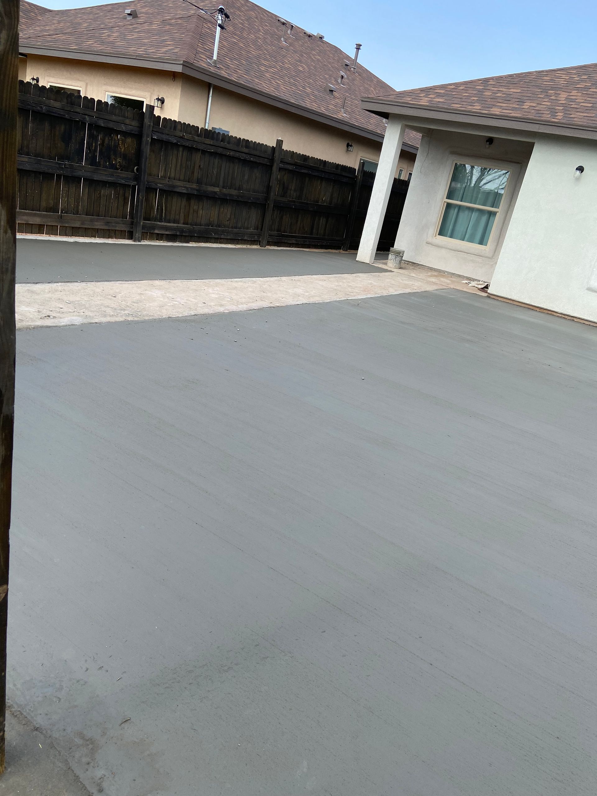 Driveway beside a house with white walls, a wooden fence, and a covered patio under a cloudy sky