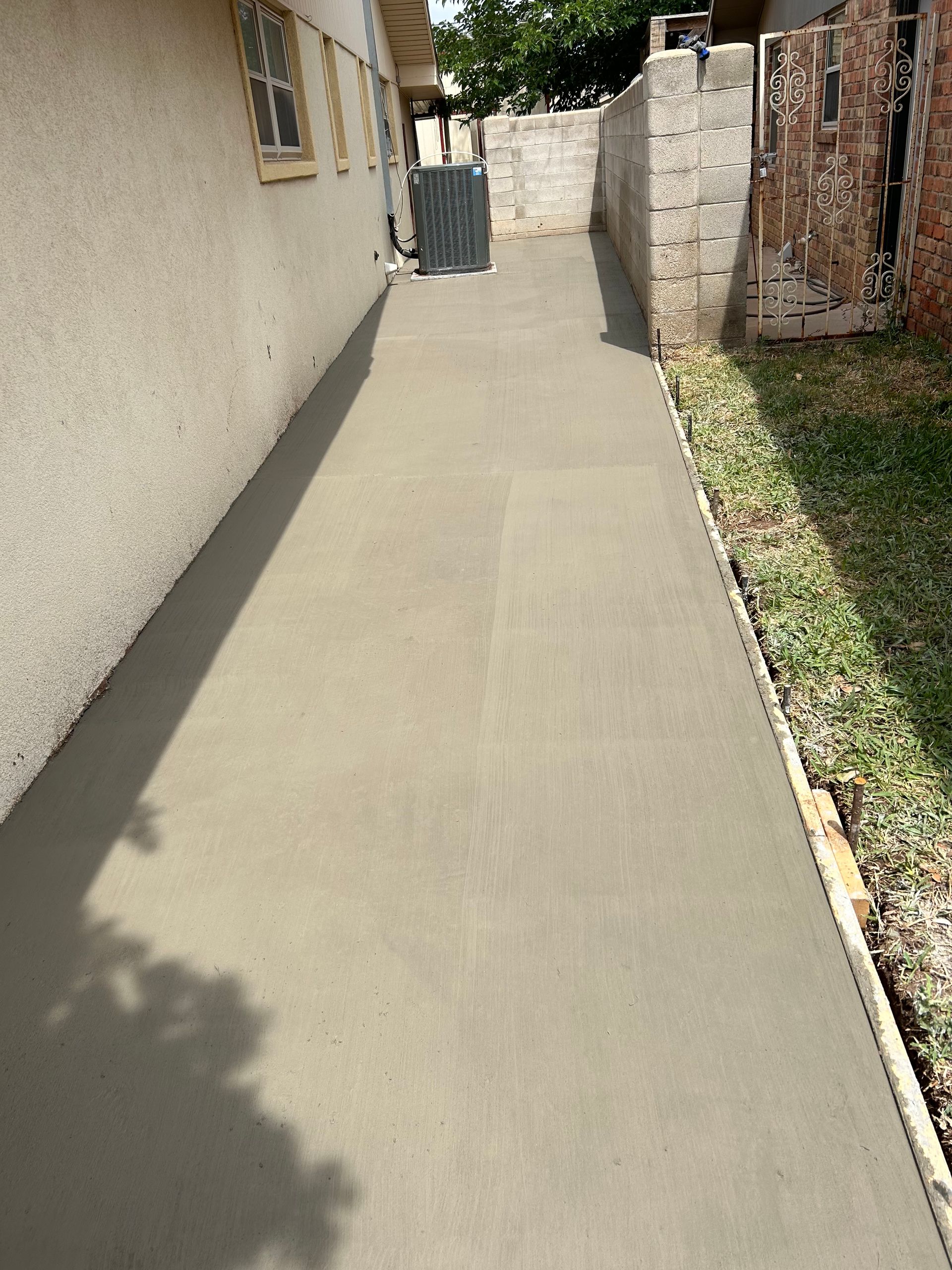 Narrow concrete walkway beside a stucco house, with a stone wall and grass on the right.