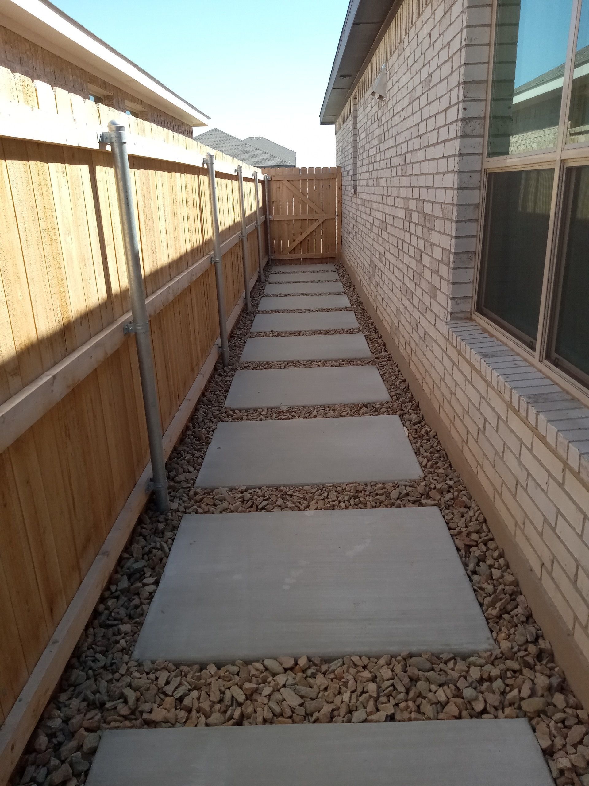 Narrow side yard with concrete stepping stones, gravel, wooden fence, and brick house wall.