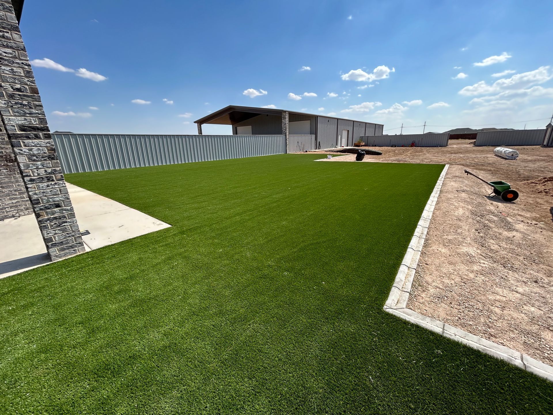 Newly landscaped green lawn beside a stone building and gravel yard under a bright blue sky.