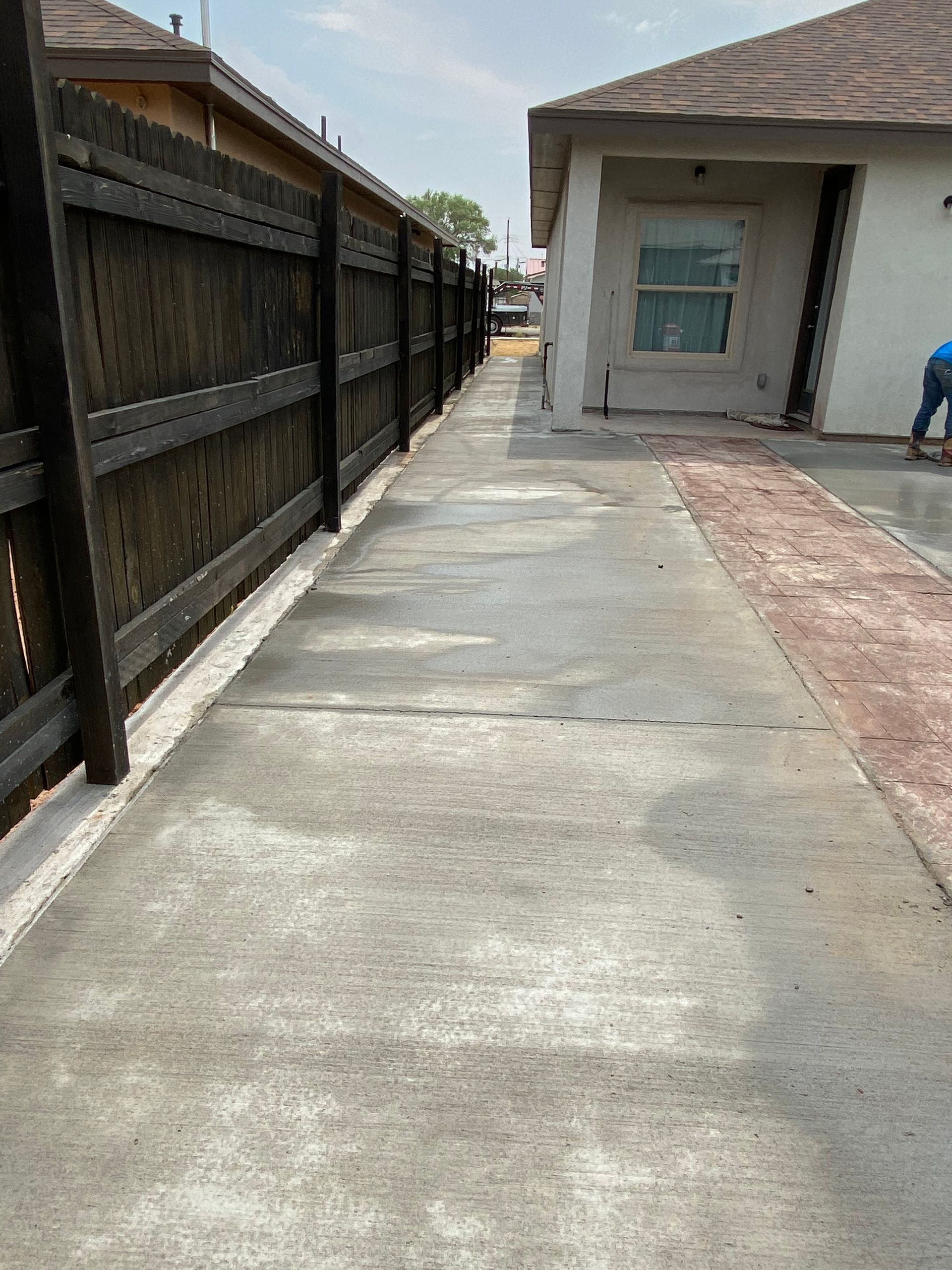 Narrow concrete walkway beside a house and wooden fence, with a person working near the doorway.