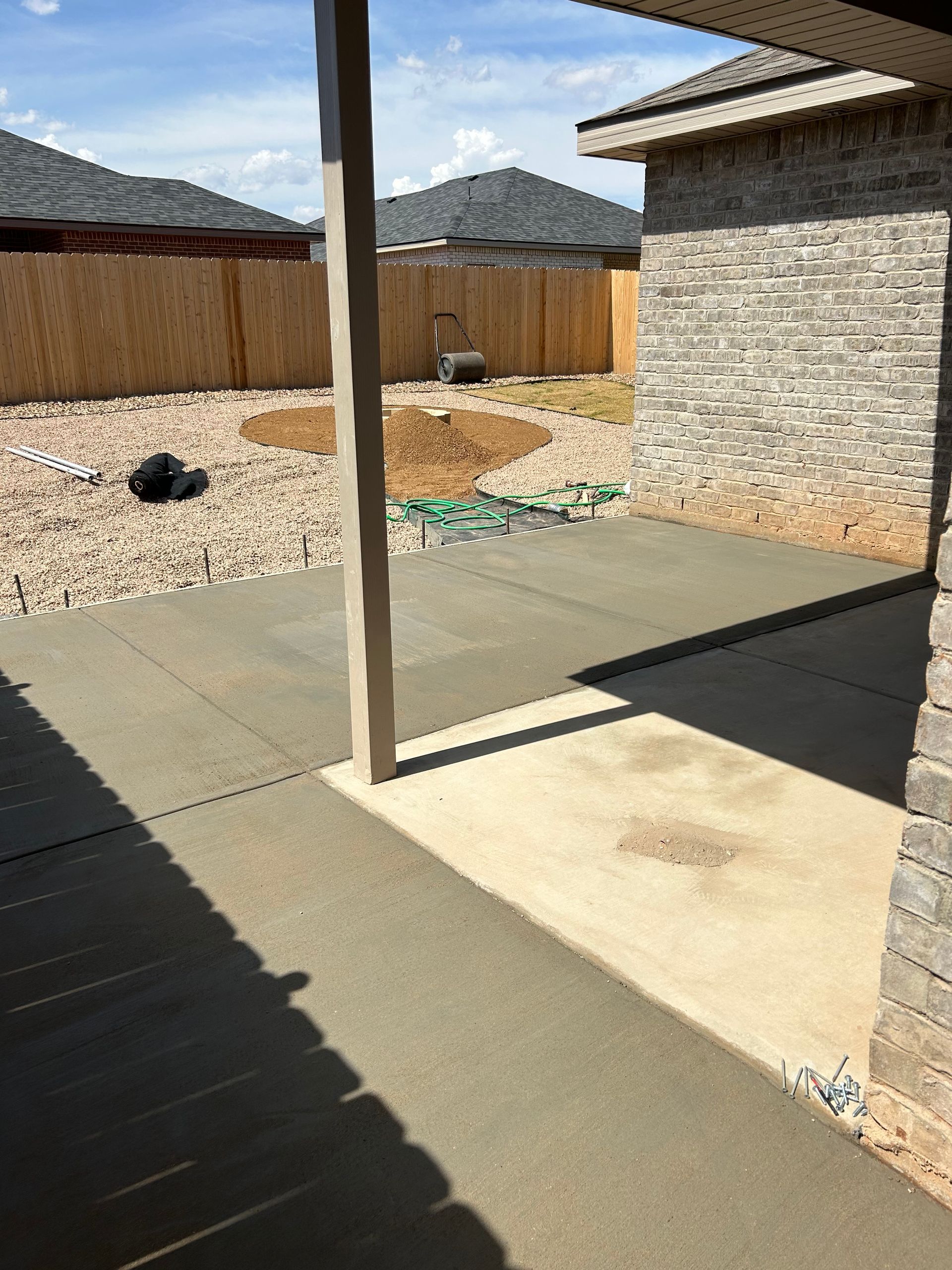 Backyard patio with concrete slab, wooden fence, stone house wall, and a tall post casting shadows.