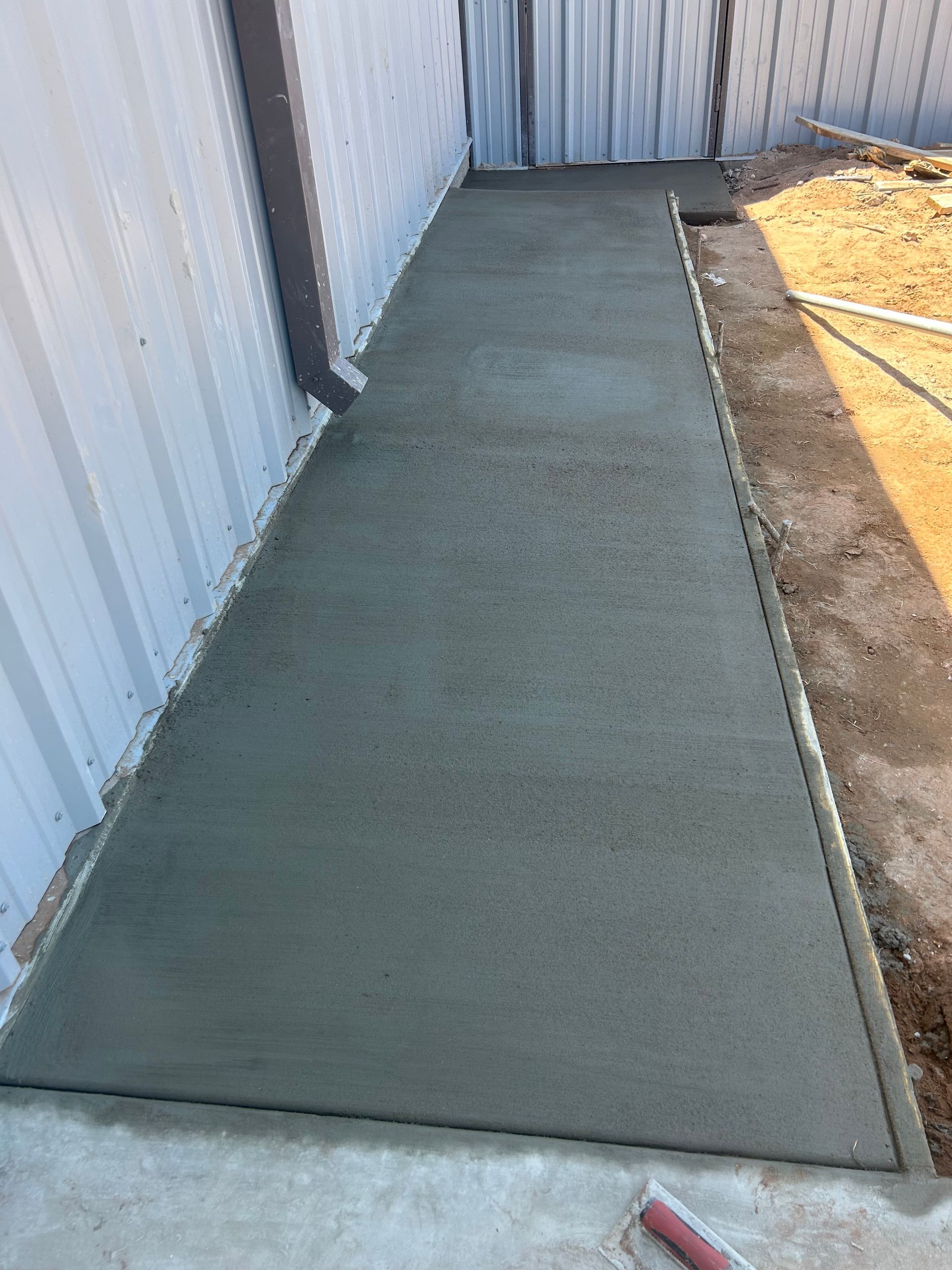 Concrete walkway beside a metal building, bordered by dirt and a hose.