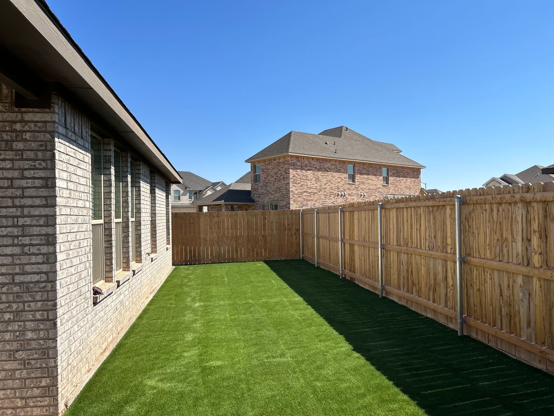 Narrow fenced backyard with green grass, brick house on the left, and neighboring homes under a clear blue sky