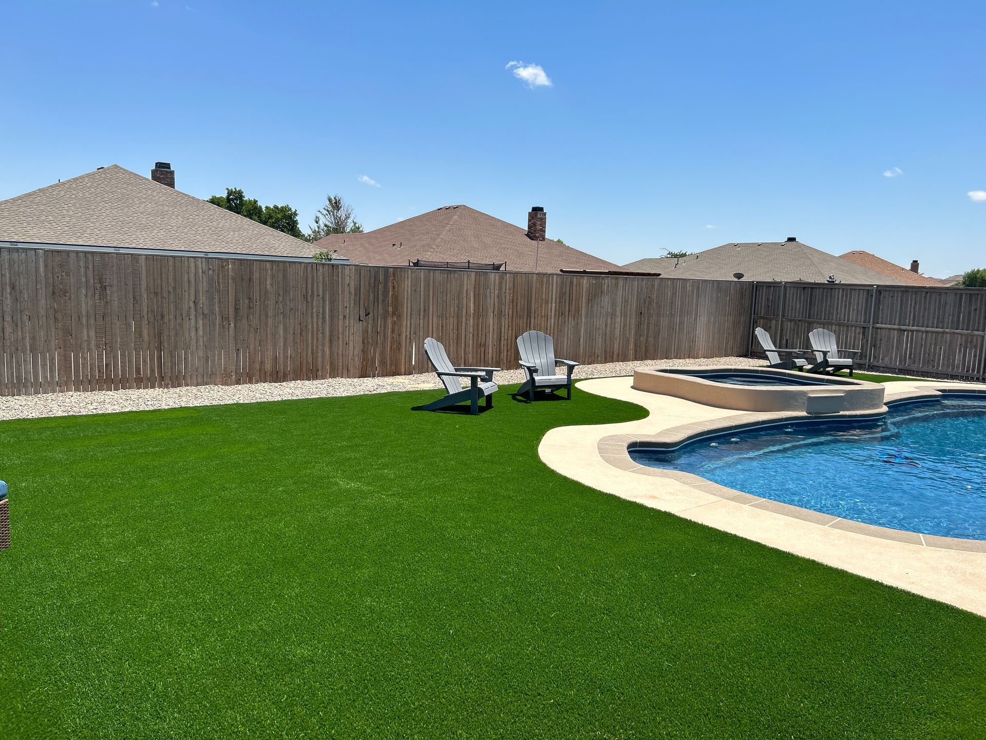 Backyard pool with green lawn, patio chairs, wooden fence, and clear blue sky