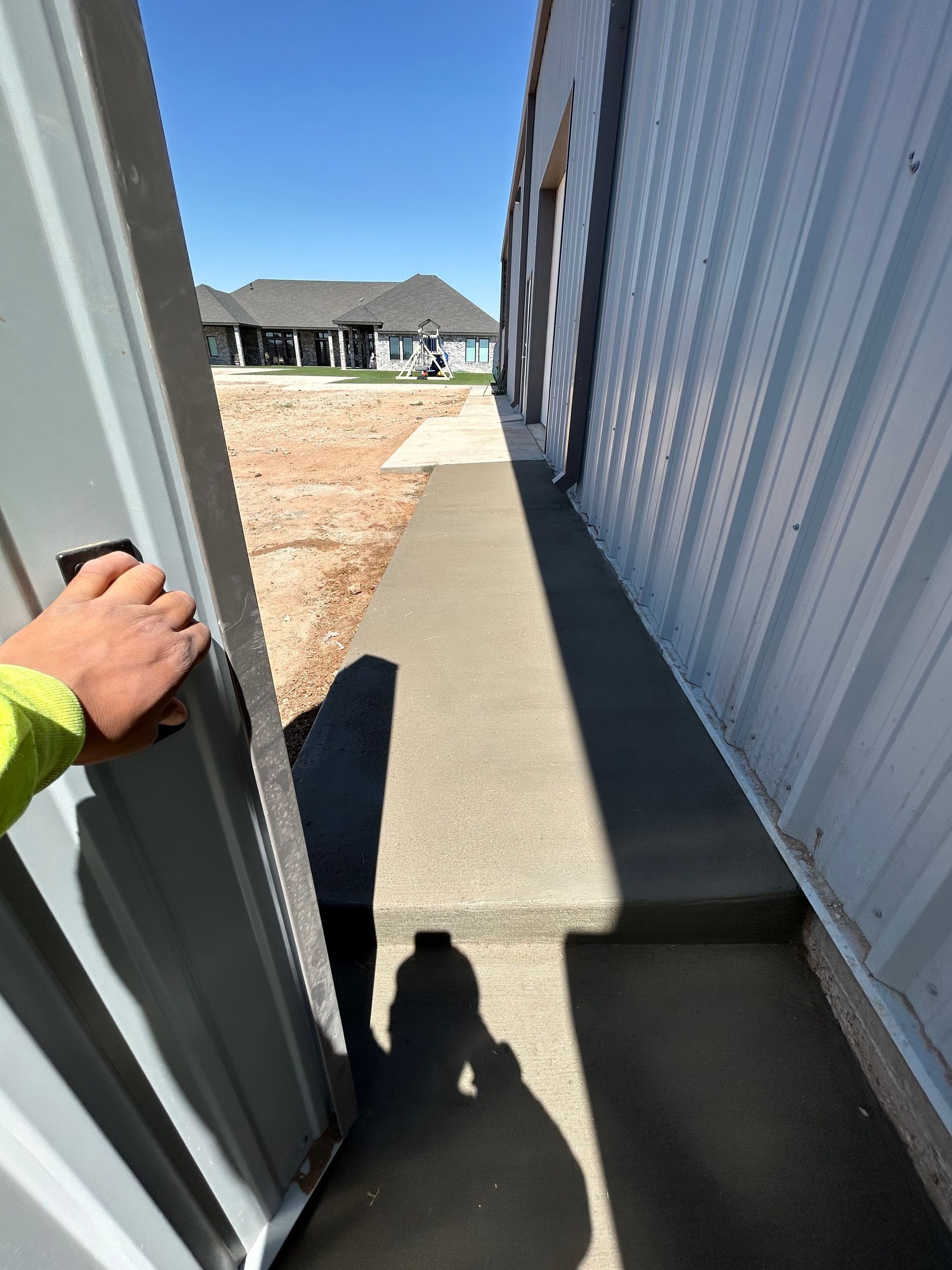 Hand on a metal door beside a narrow concrete path between buildings, with a house visible ahead.