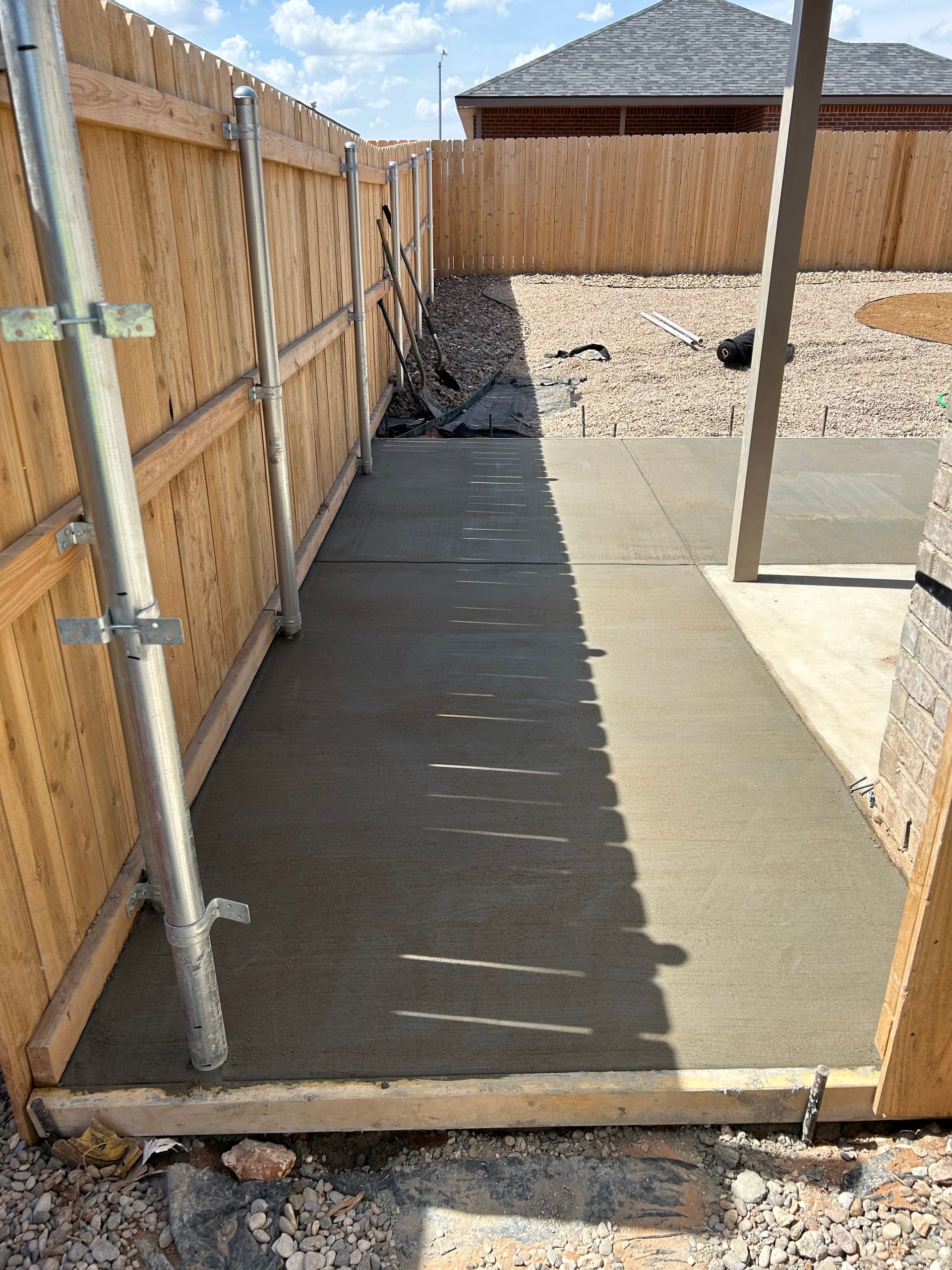 Narrow concrete walkway beside a wooden fence, ending at a gravel yard and a small covered area.