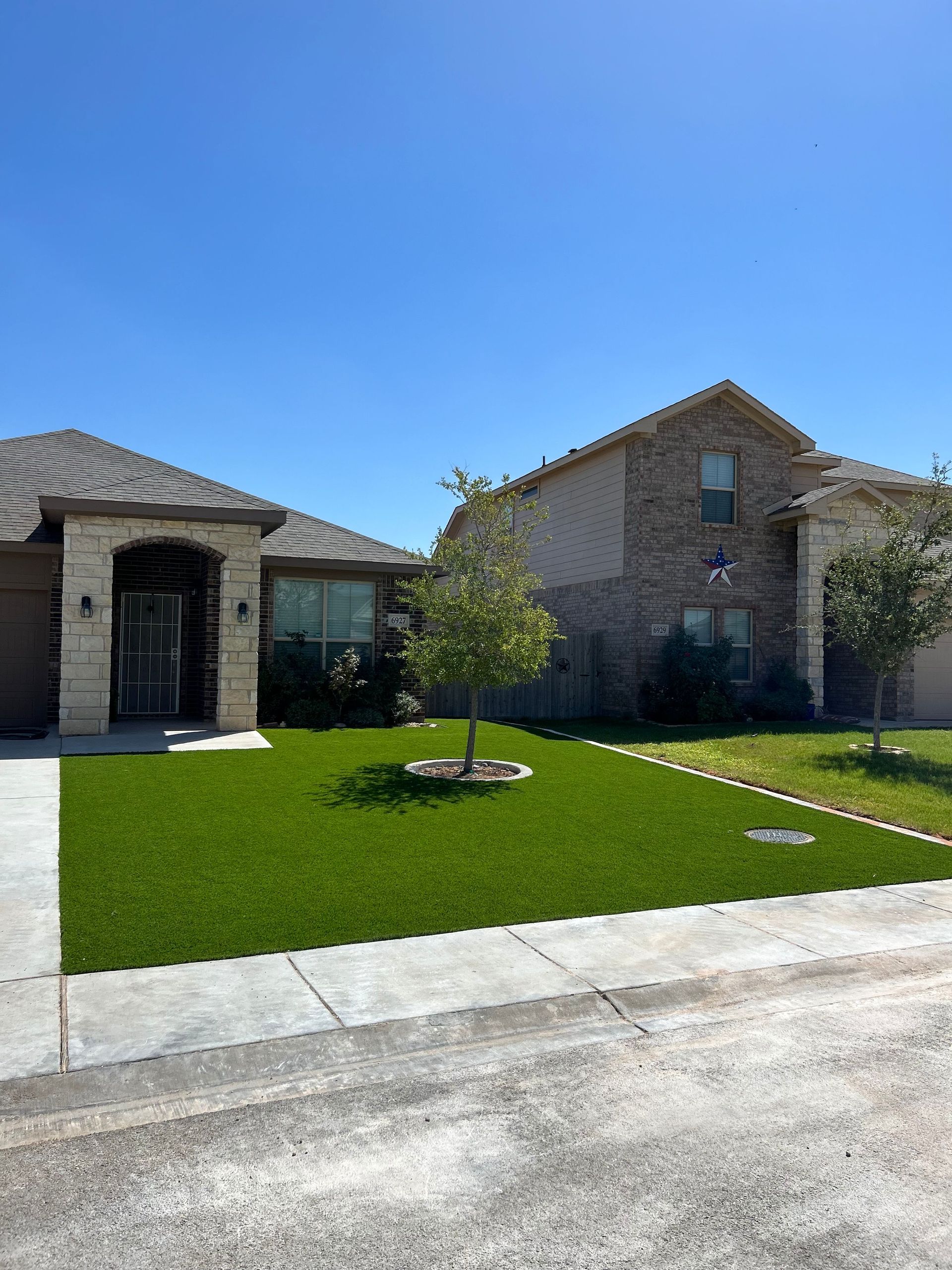 Suburban brick house with a green front lawn, small tree, and concrete driveway under a clear blue sky