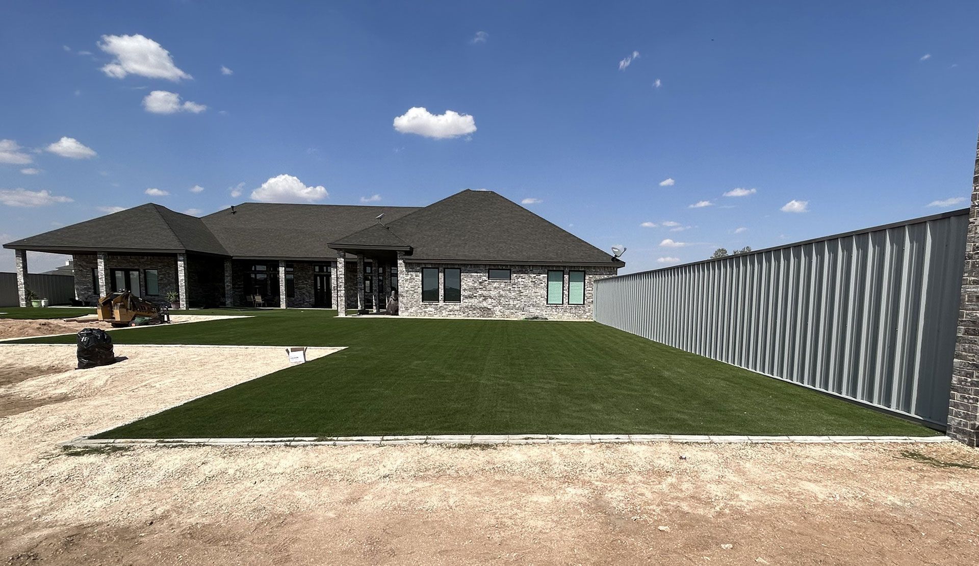 Suburban house with a large green lawn, gravel driveway, and a tall metal fence under a blue sky