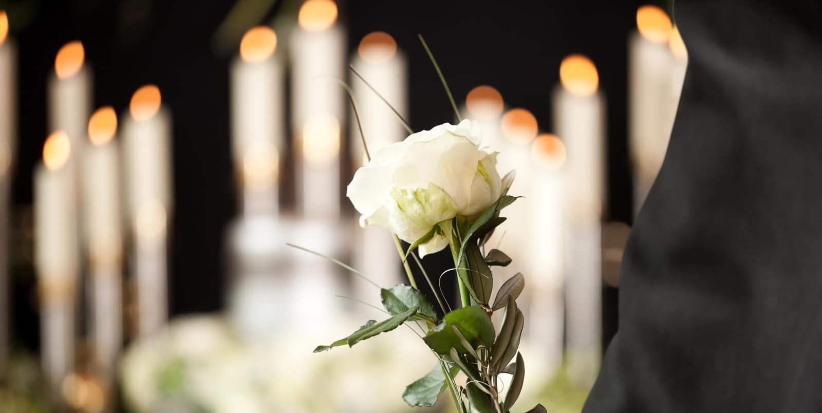 A man is holding a white rose in front of candles at a funeral.