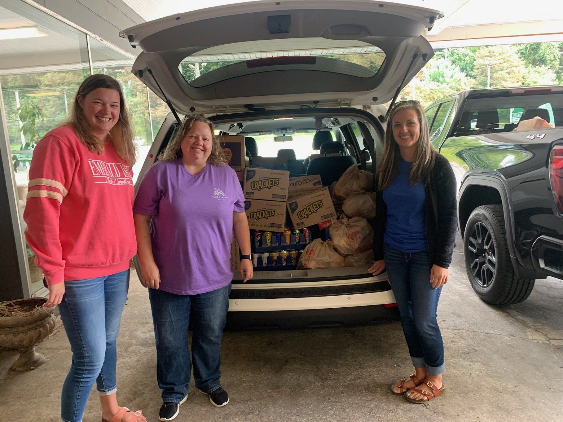 Three women are standing in front of a car with boxes in the back.