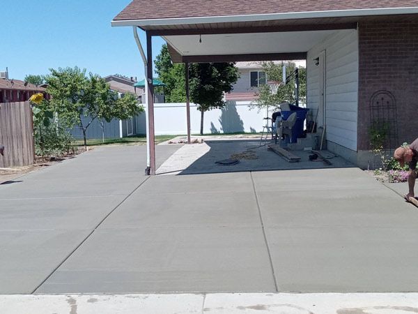 Driveway beside a house with a carport, concrete path, and blue trash bins in the shade.