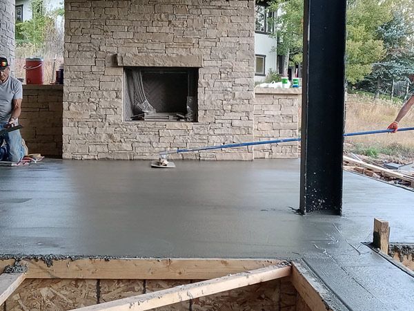 Freshly poured concrete patio around a stone fireplace, with construction tools and a worker at left.