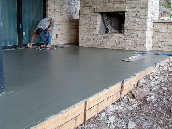Worker smoothing a concrete patio slab beside a stone fireplace and house exterior