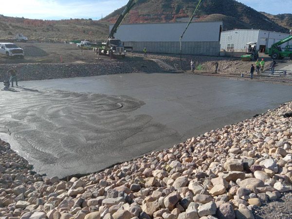 Workers smooth wet concrete at a construction site beside a rocky embankment and hills.