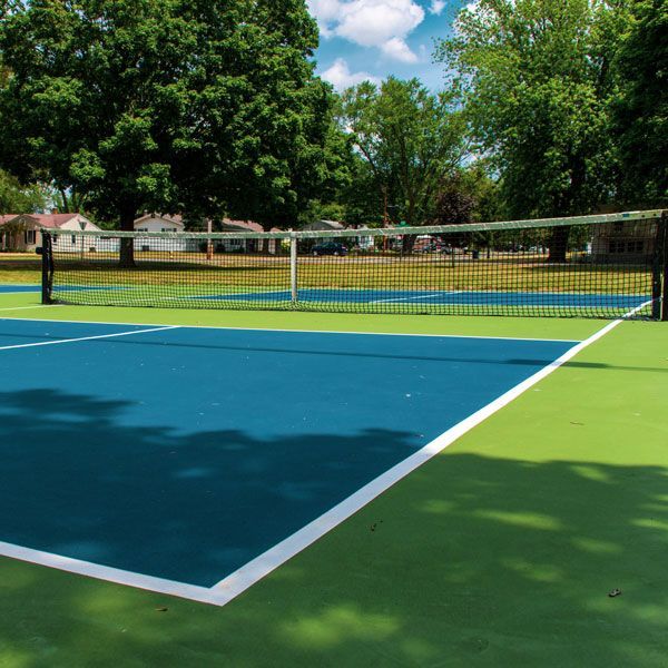 Empty outdoor tennis court with blue and green surface, net centered, under trees on a sunny day