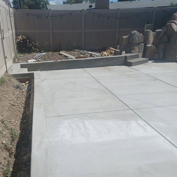 Concrete patio with a low retaining wall, bordered by dirt and a wooden fence in a backyard.