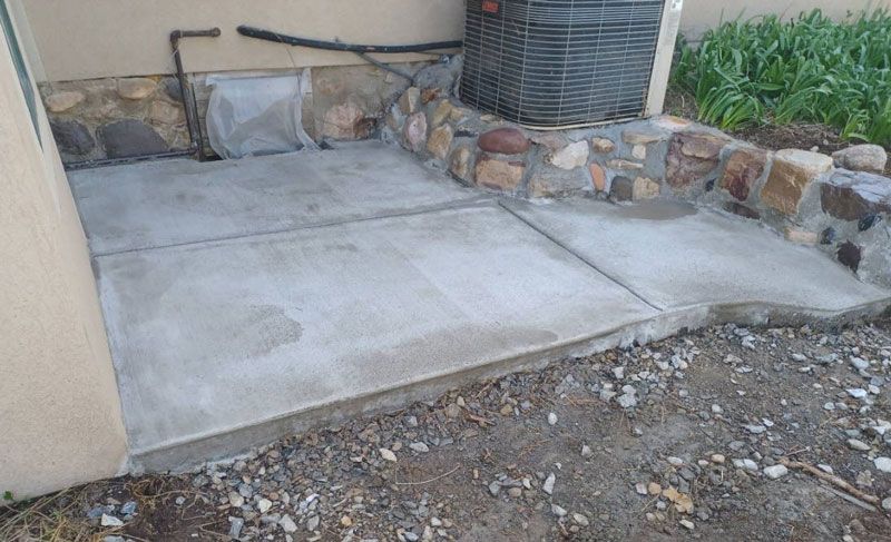 Concrete patio next to a stone retaining wall and gravel yard with outdoor HVAC equipment