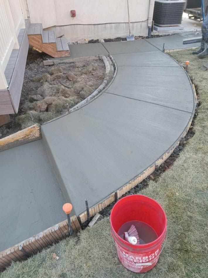 Curved concrete walkway beside a house, with a red bucket in the foreground and bare landscaping along the edge.