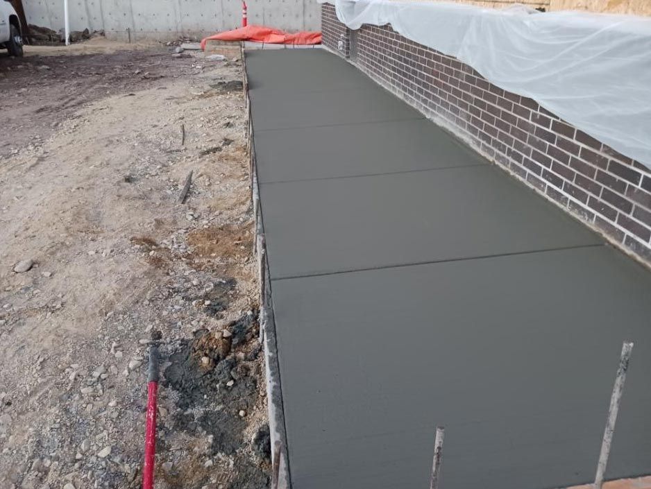 Freshly poured gray concrete walkway beside a brick house, with plastic sheeting above the wall.