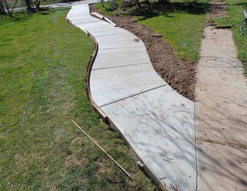Curving concrete sidewalk under construction beside a grassy yard and dirt trench
